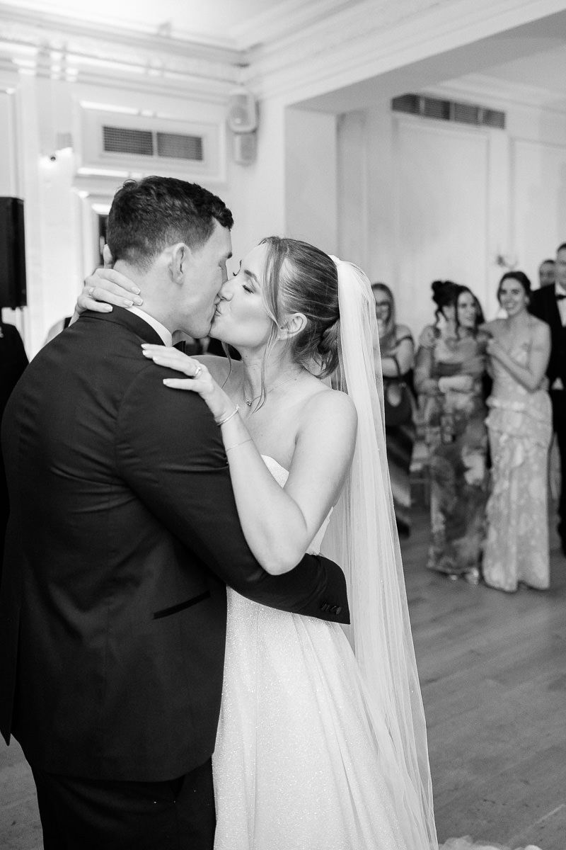 A bride in a white gown and veil shares a romantic first kiss with her groom in a black tuxedo as guests look on.