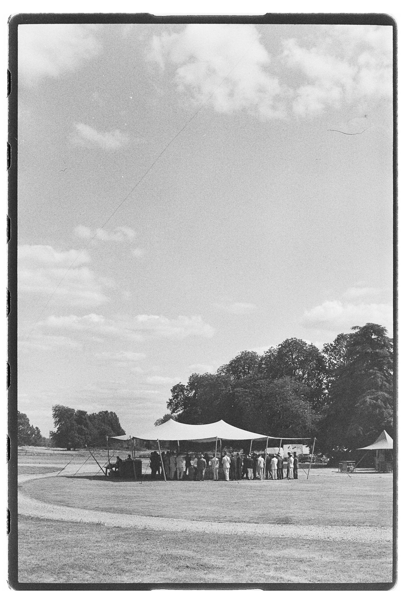 Black and white photo of people gathered under a large tent on an open field with trees in the background on a sunny day.