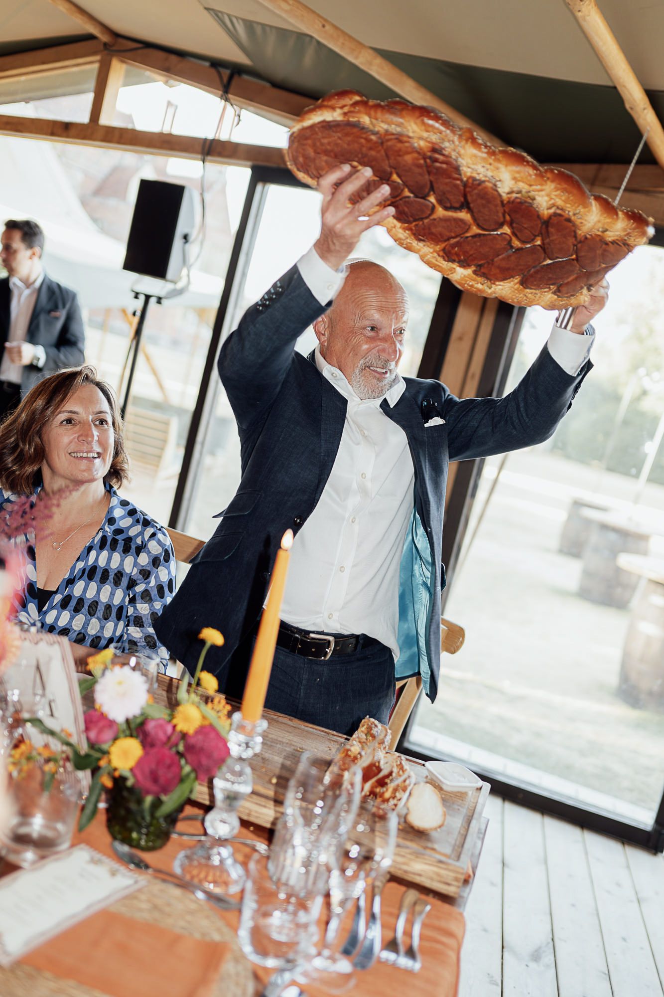 Man in navy blazer proudly holds up large braided bread at elegant decorated dining table during celebration event.