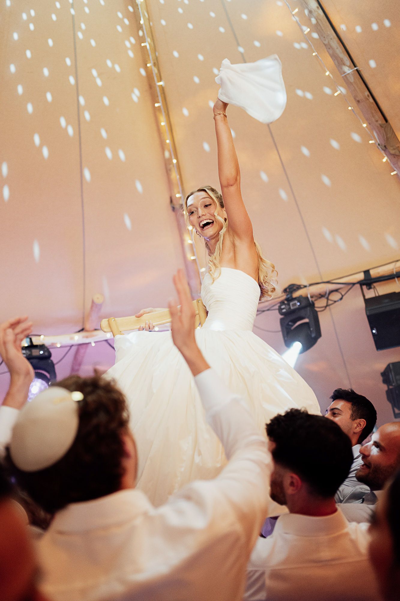 Joyful bride lifted by guests at Jewish wedding reception, raising napkin in celebration under festive lights.