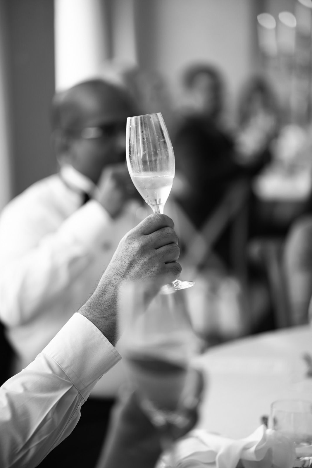A hand raises a champagne flute in a toast at an elegant wedding reception in black and white.