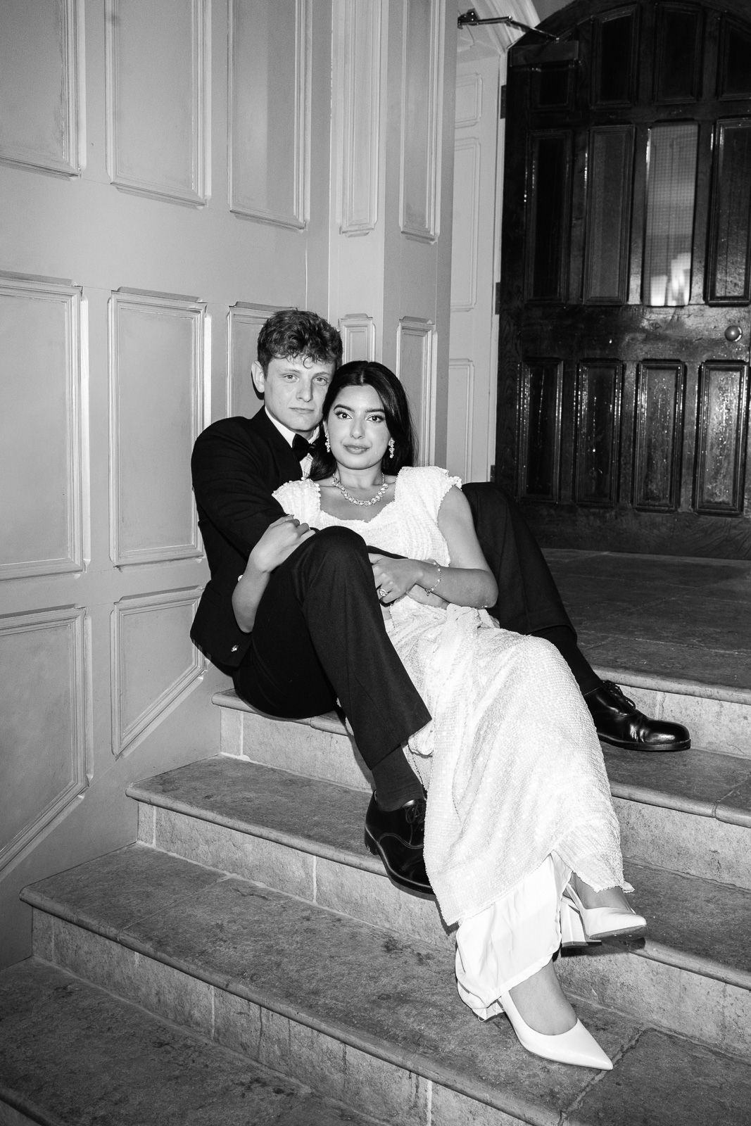 Black and white photo of newlyweds sitting together on stone steps outside an elegant venue door.