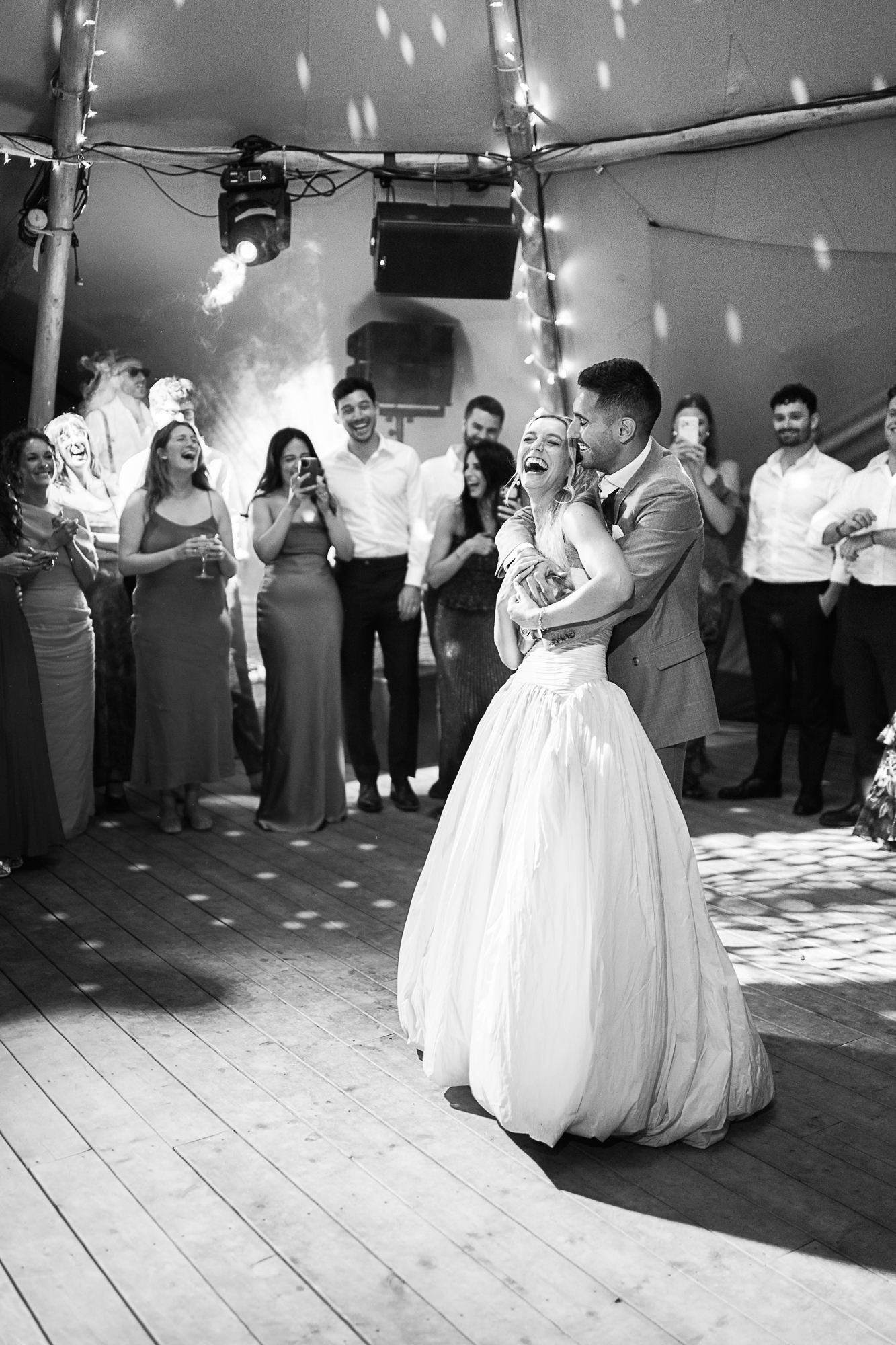 Black and white photo of bride and groom sharing first dance surrounded by cheering wedding guests under fairy lights.