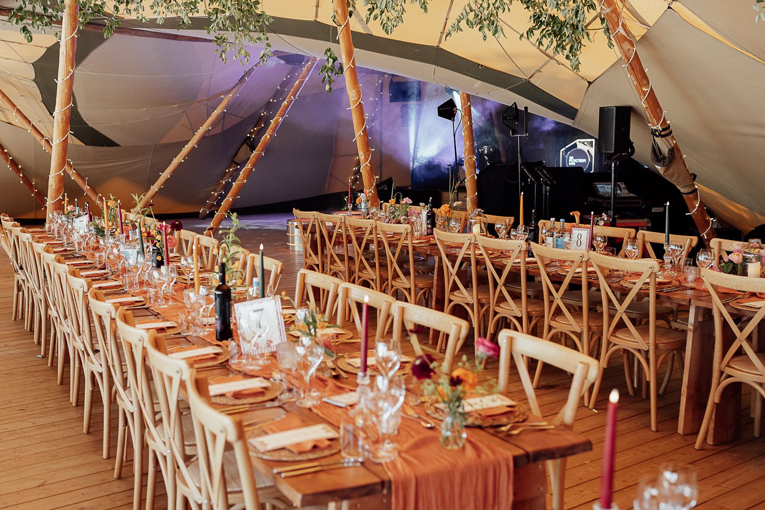 Elegant tipi wedding reception with long wooden tables, terracotta runners, colorful florals, and string lights.