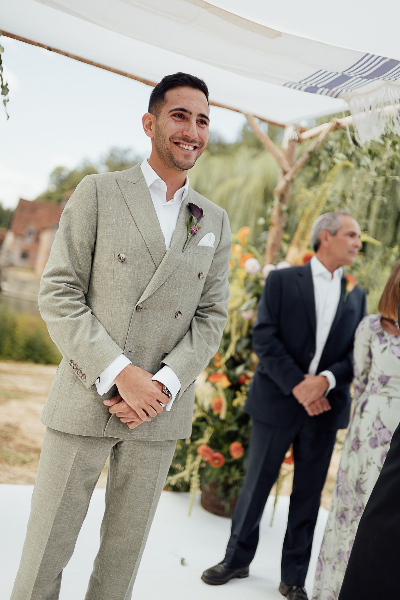 Smiling groom in light gray double-breasted suit with floral boutonniere standing at outdoor wedding ceremony altar.
