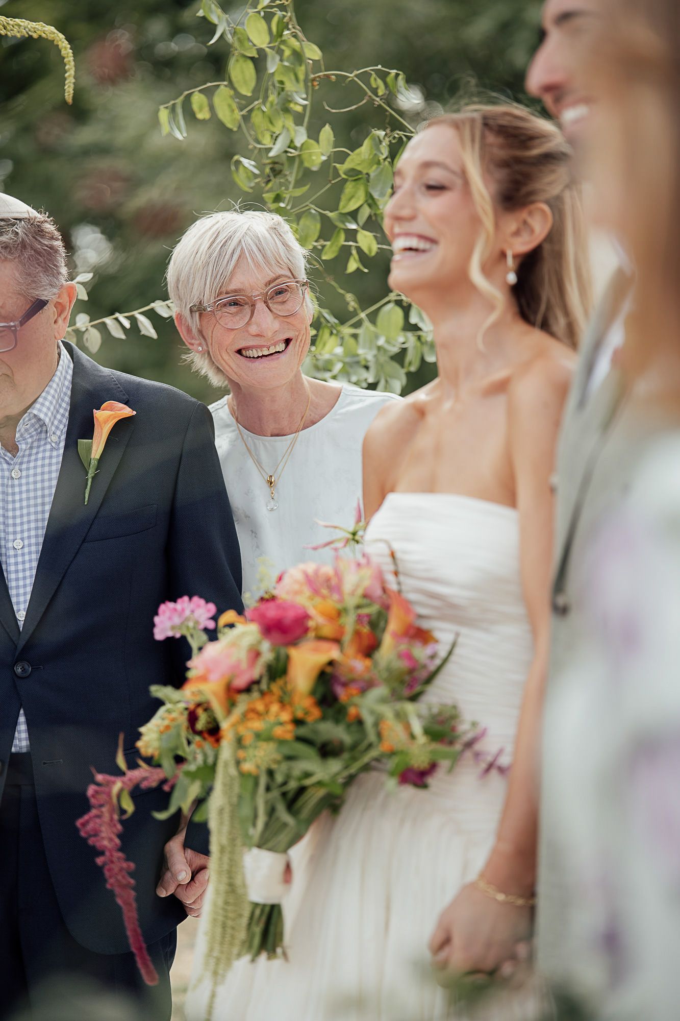 Smiling bride in white dress holds colorful bouquet walking with elderly woman in dark suit at outdoor wedding ceremony.