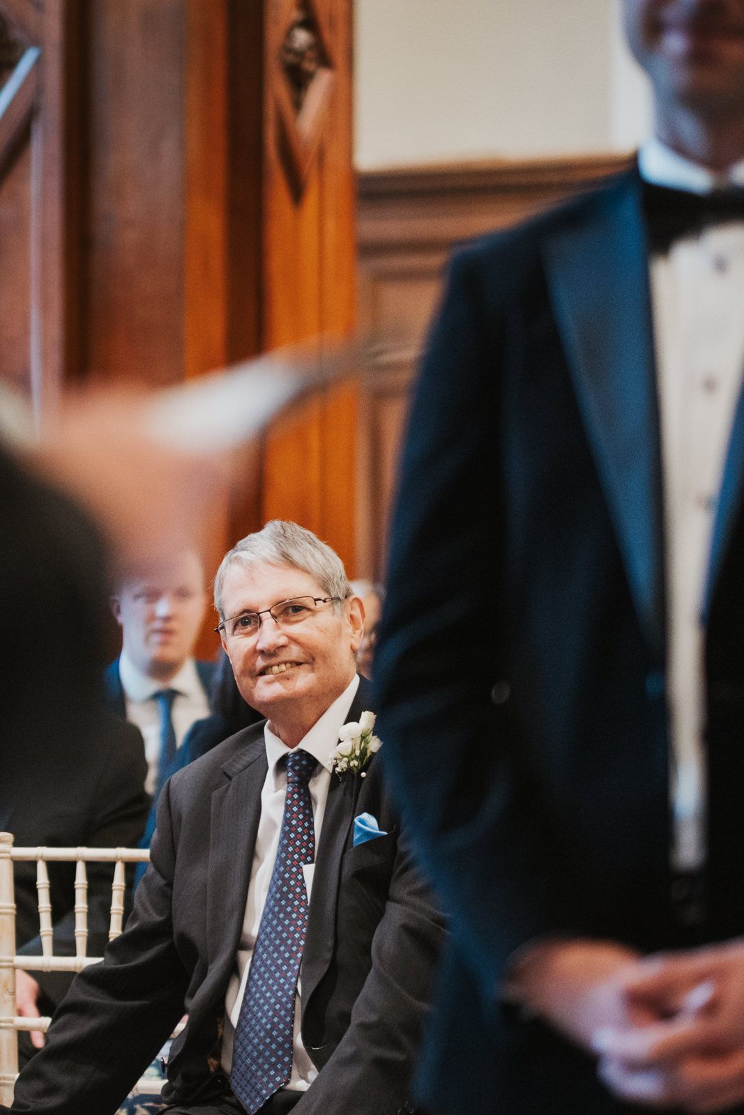 Smiling wedding guest dressed in dark suit with polka dot tie watches ceremony from wooden chair.