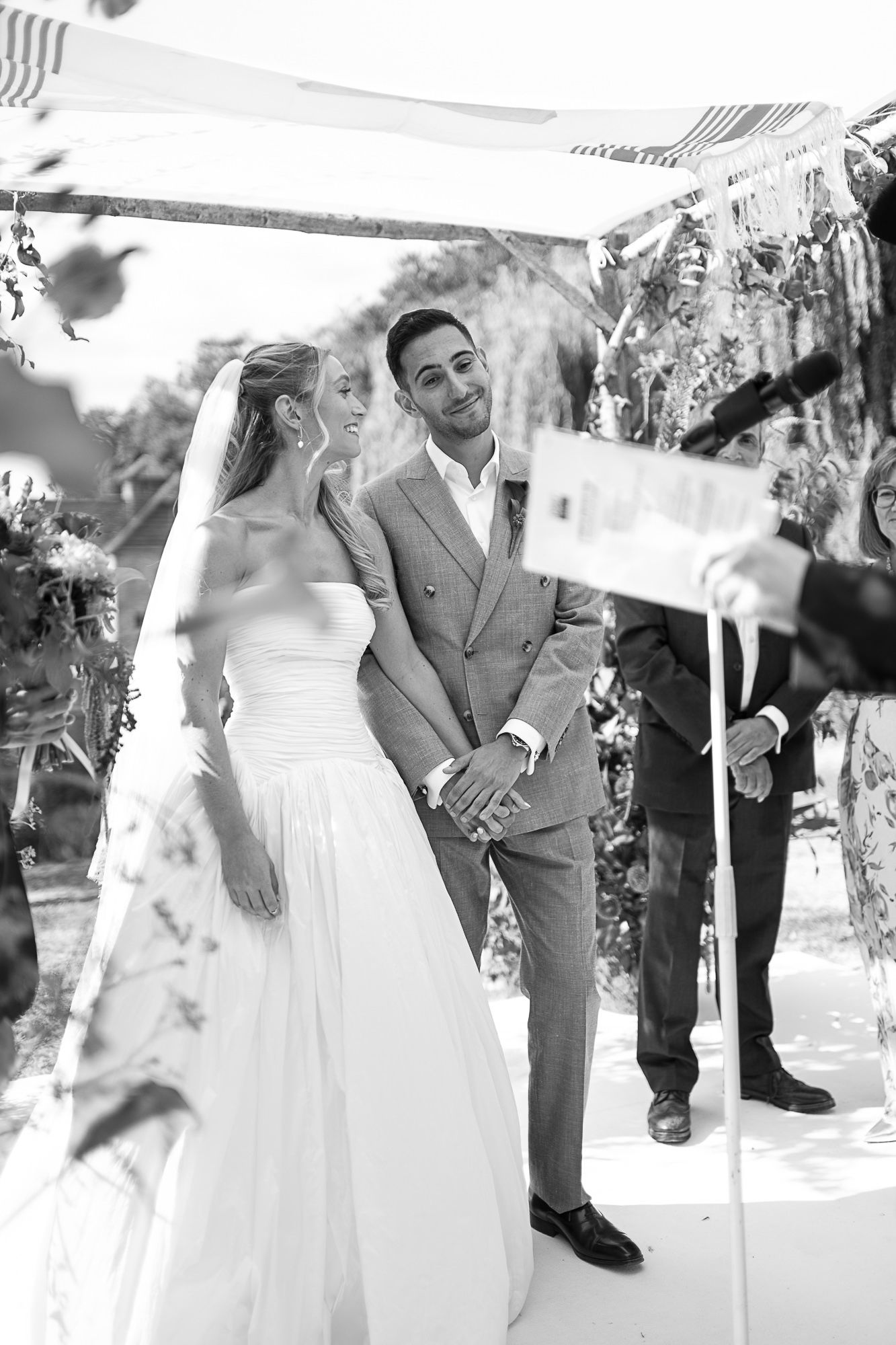 Black and white photo of smiling bride and groom holding hands at outdoor wedding ceremony under floral arch.