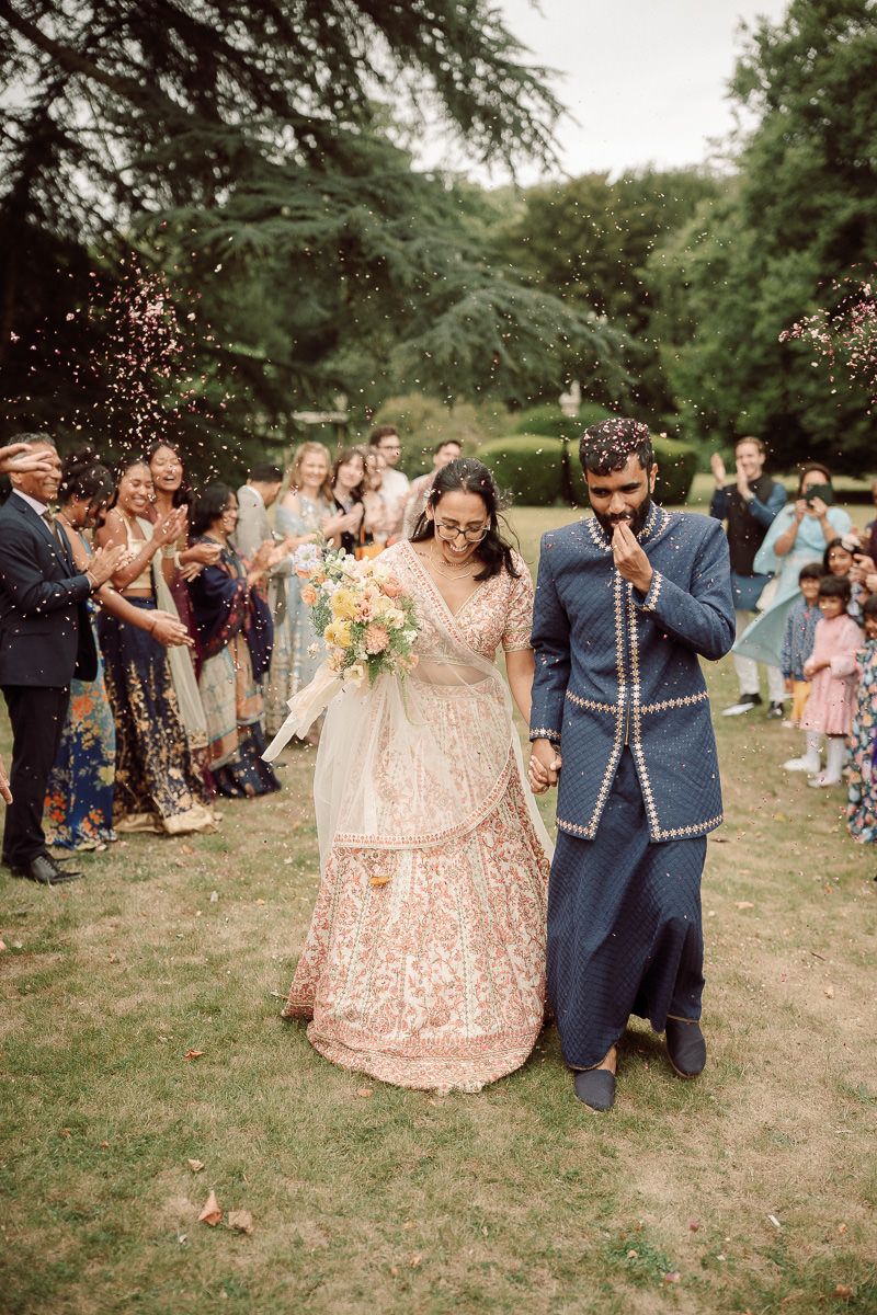 Wedding guests toss confetti at a couple in traditional Indian fusion attire during an outdoor celebration.