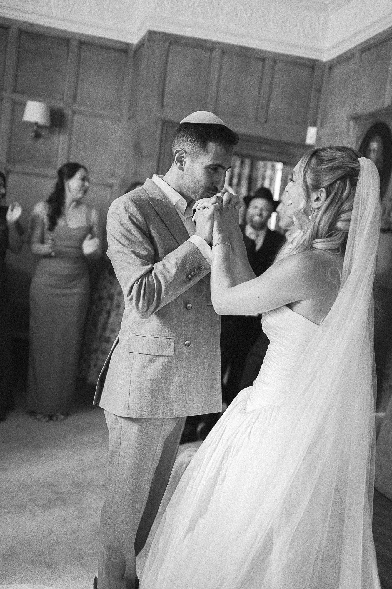 Black and white photo of bride and groom sharing first dance, groom wearing kippah, guests watching in background.