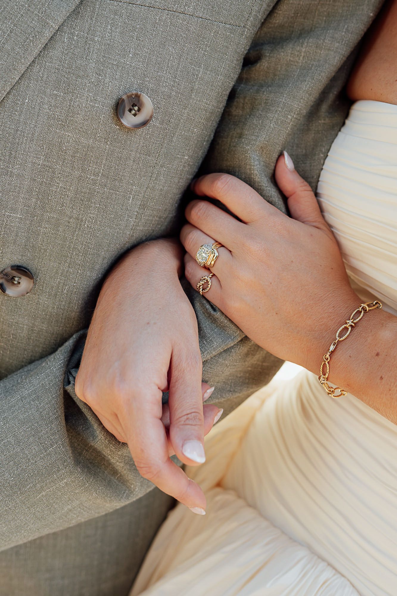 Bride wearing a gold diamond ring and chain bracelet holds groom's arm in gray suit at wedding.