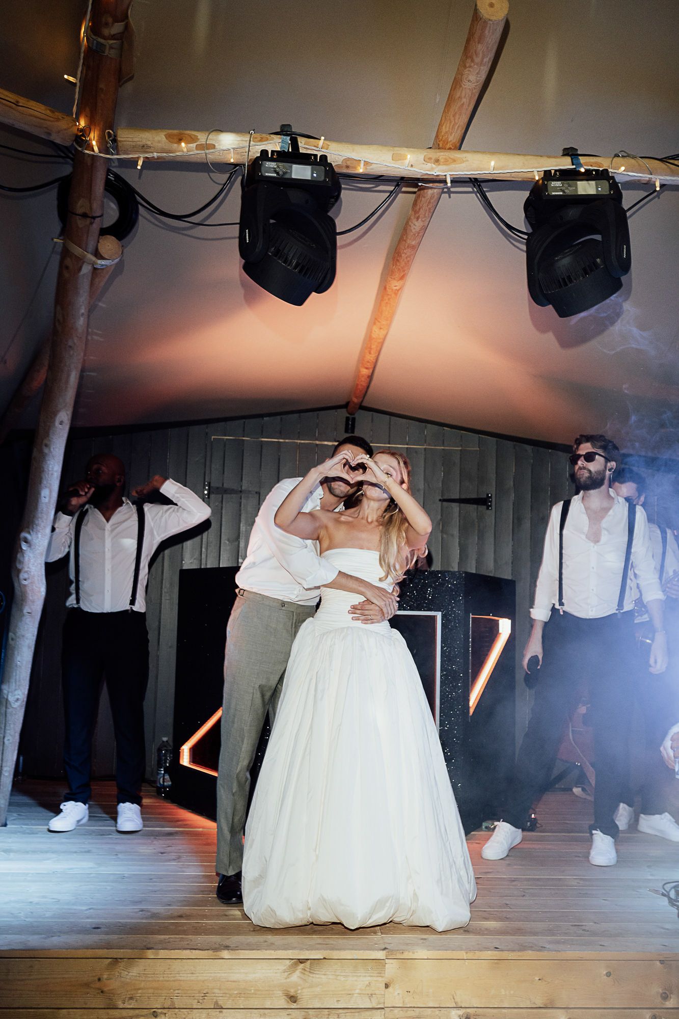 Bride and groom share first dance making heart shape with hands at elegant wedding reception with stage lighting.