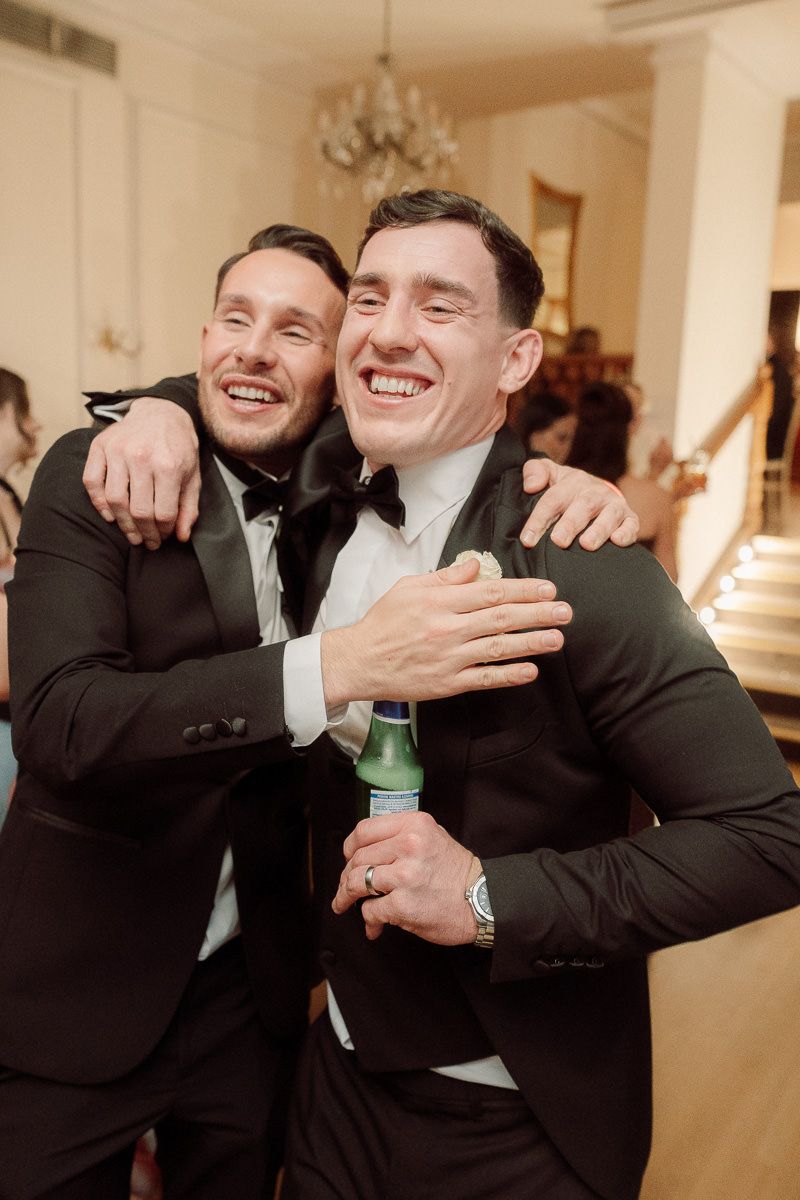 Two people in black suits embrace and laugh together at a formal event.