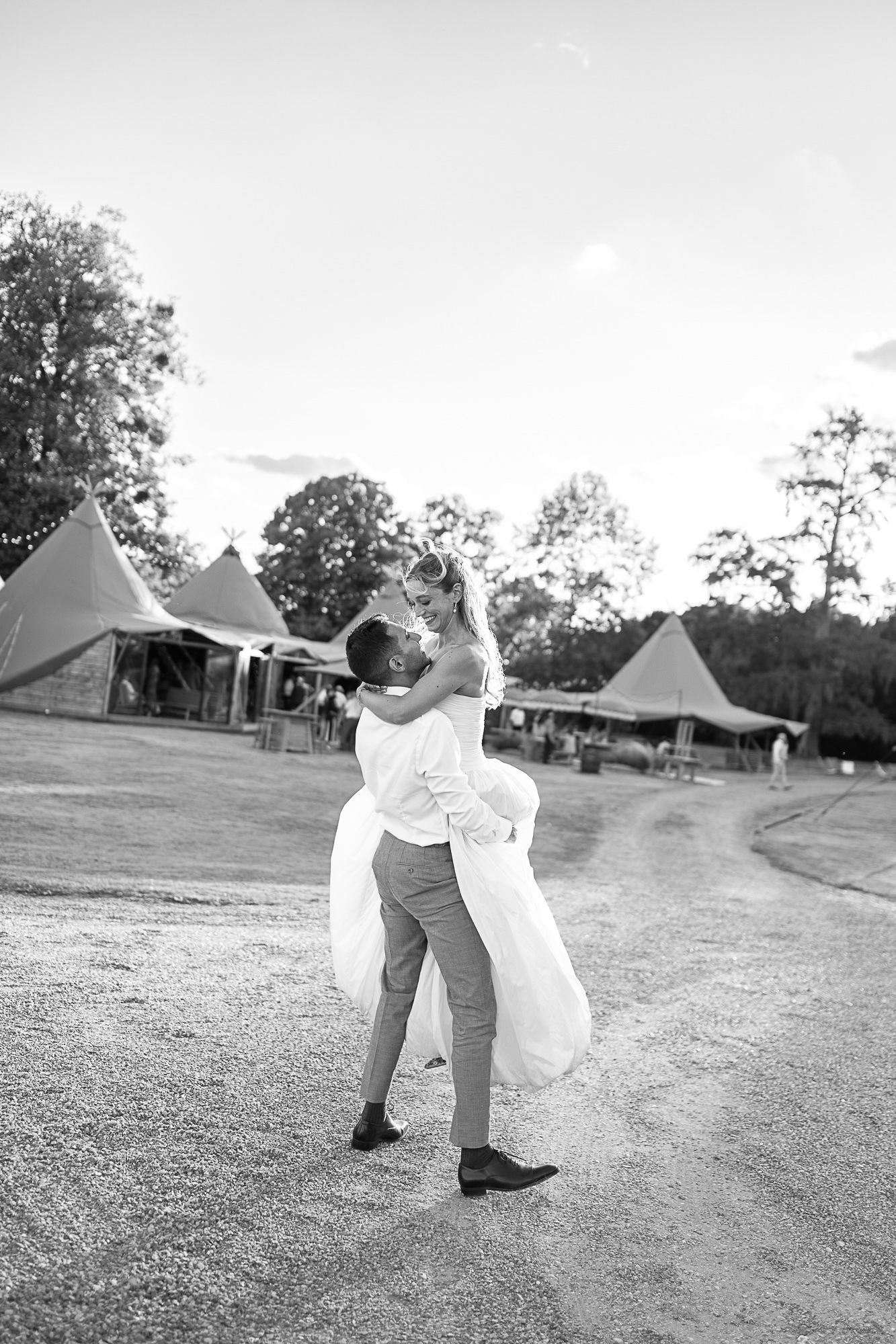Groom lifts bride in wedding dress at outdoor tipi venue, black and white romantic wedding photography.
