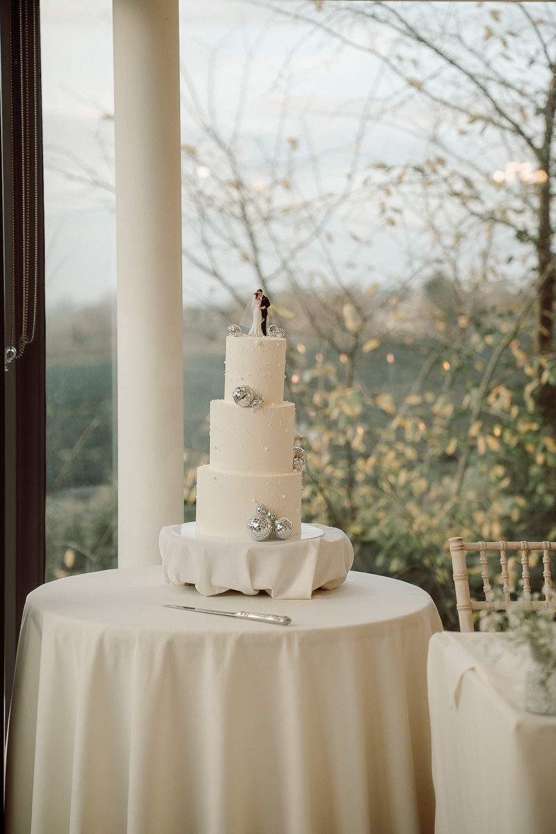Elegant white wedding cake displayed on round table with white tablecloth near large windows overlooking autumn trees.