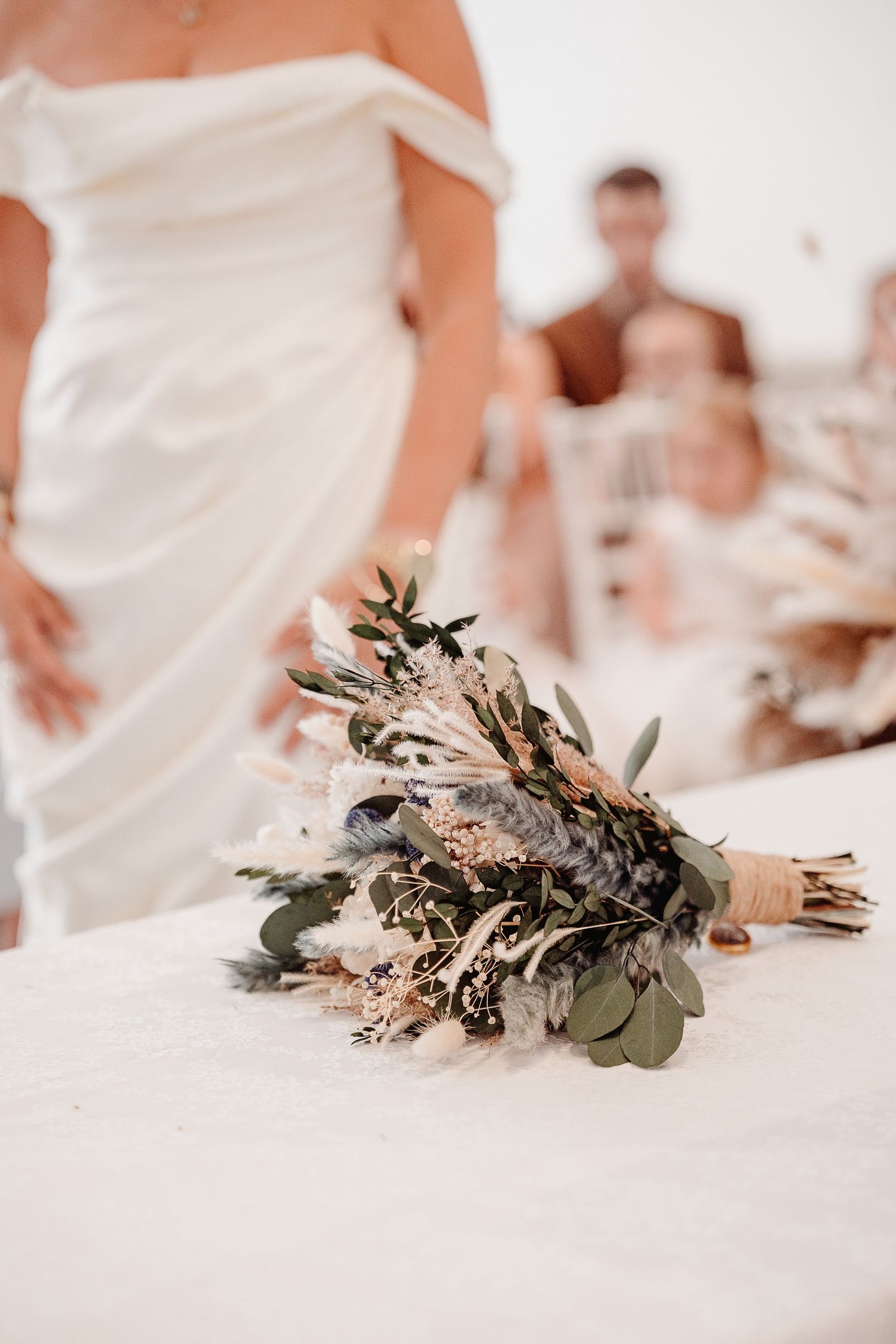 Wedding bouquet arranged with eucalyptus leaves and dried flowers lies on white table cloth in series of detail shots.