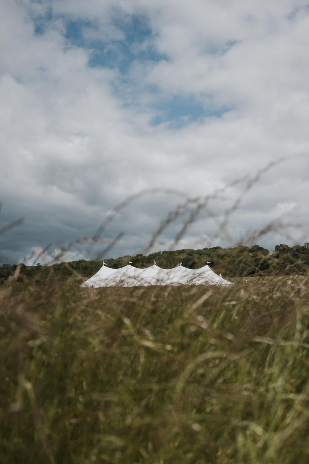 White event tent peaks visible across a field of tall grass under cloudy skies with patches of blue.