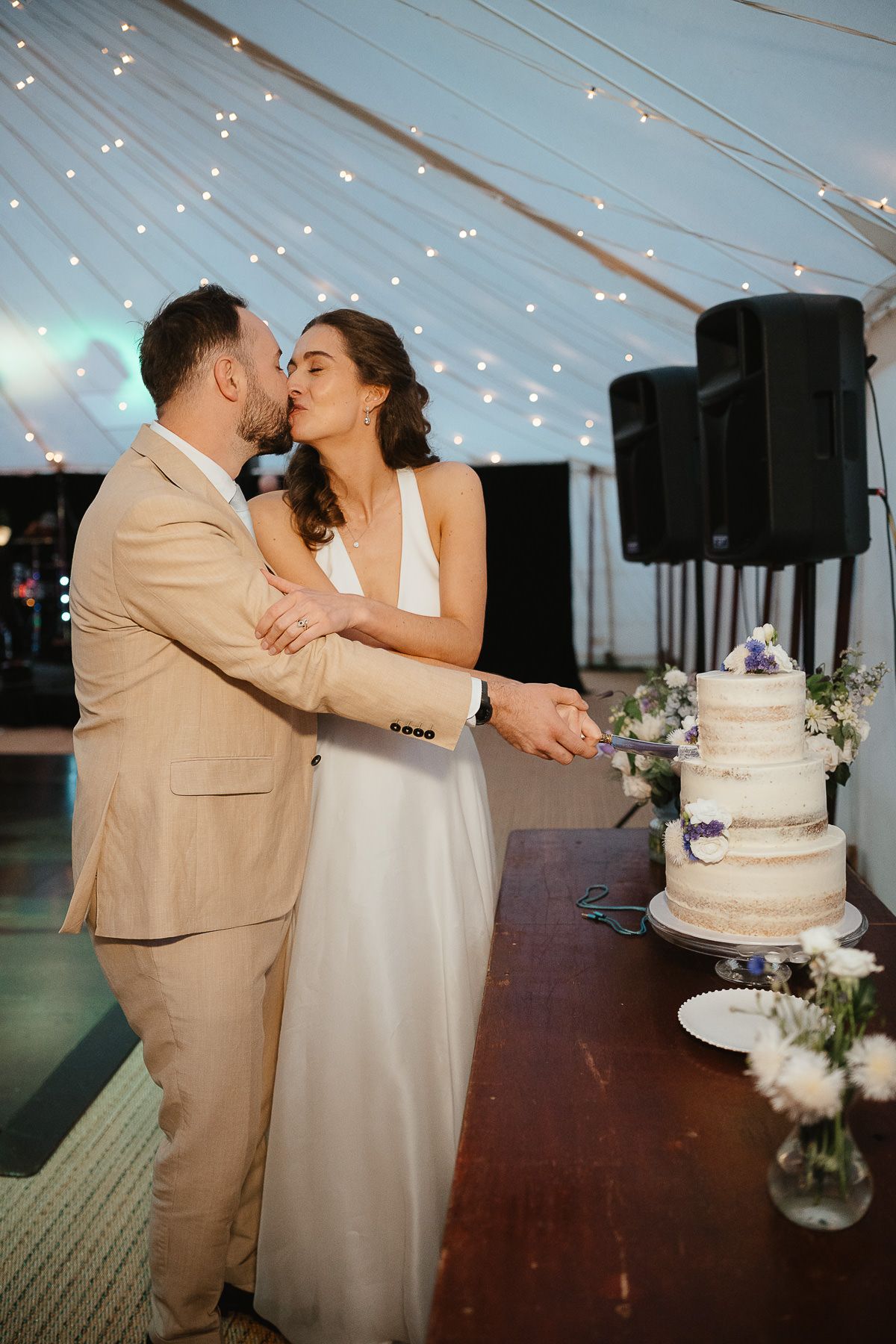 A wedding couple shares their first kiss next to their elegant white wedding cake under a tent with string lights.