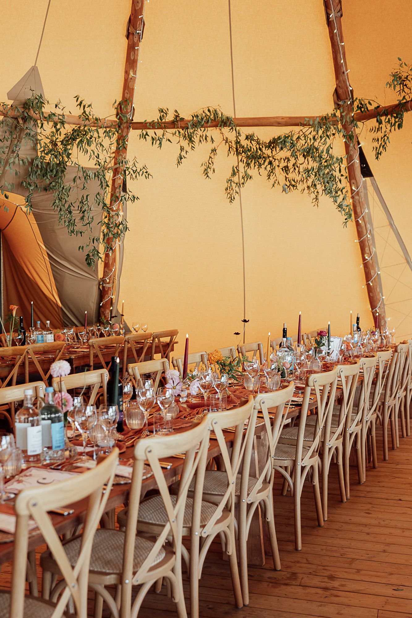 Elegant tipi wedding reception with long dining tables, white cross-back chairs, floral garlands, and warm golden tent interior.