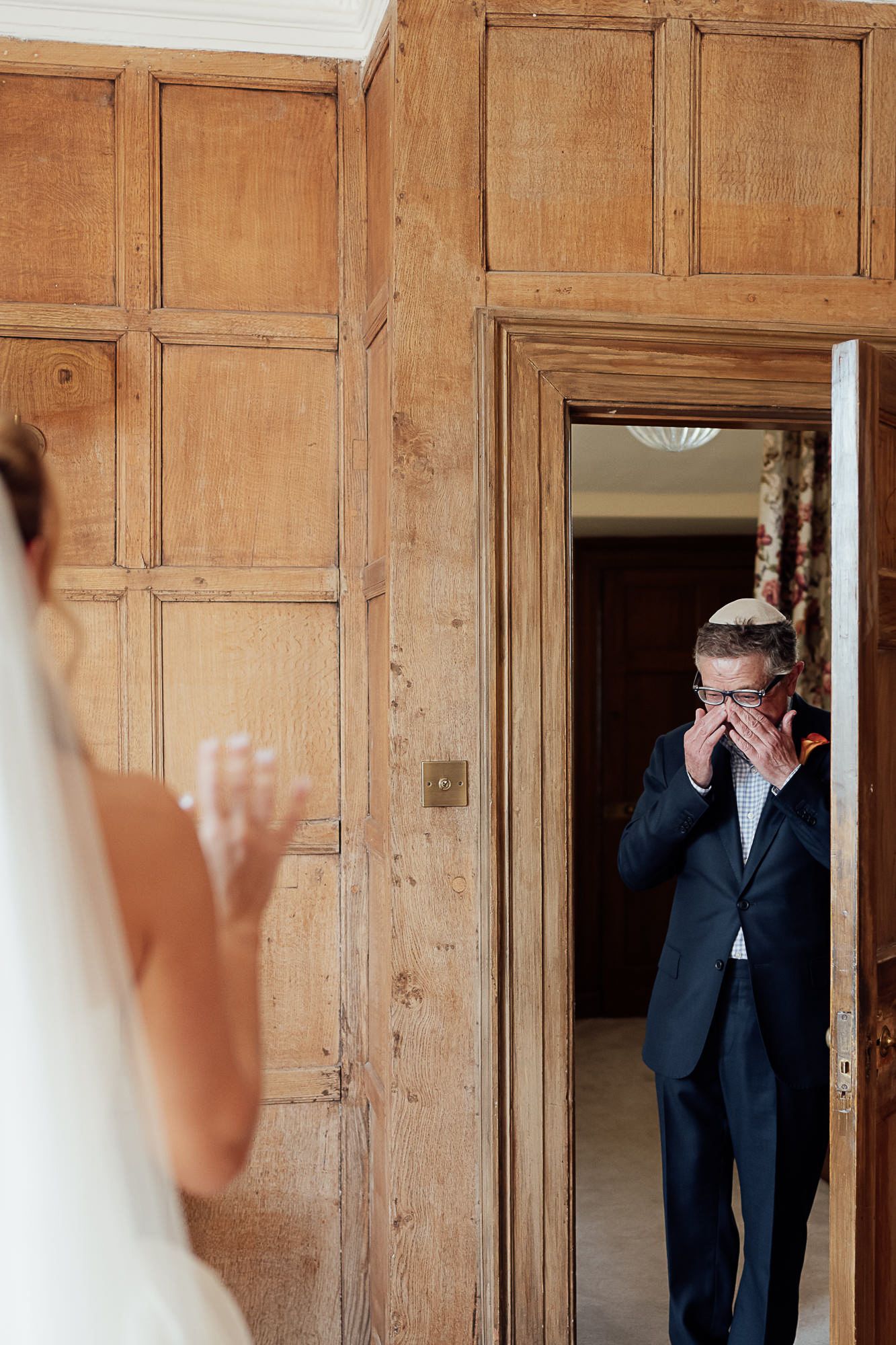 Emotional groom wiping tears seeing bride for first time in elegant wood-paneled room during wedding first look.
