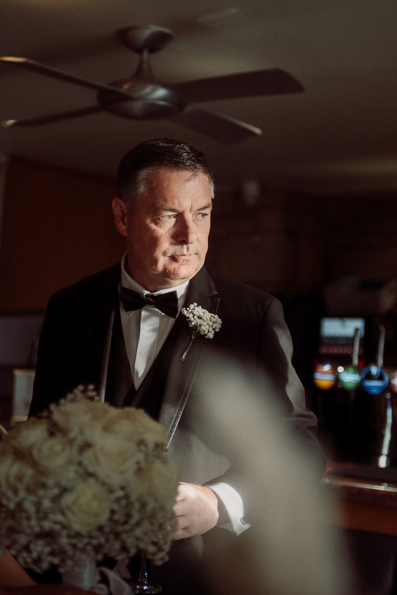 A groom in a black tuxedo smells his boutonniere in a dimly lit indoor setting.