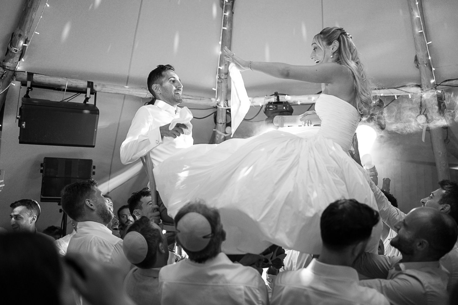 Black and white photo of bride and groom being lifted by guests during a joyful wedding reception celebration.