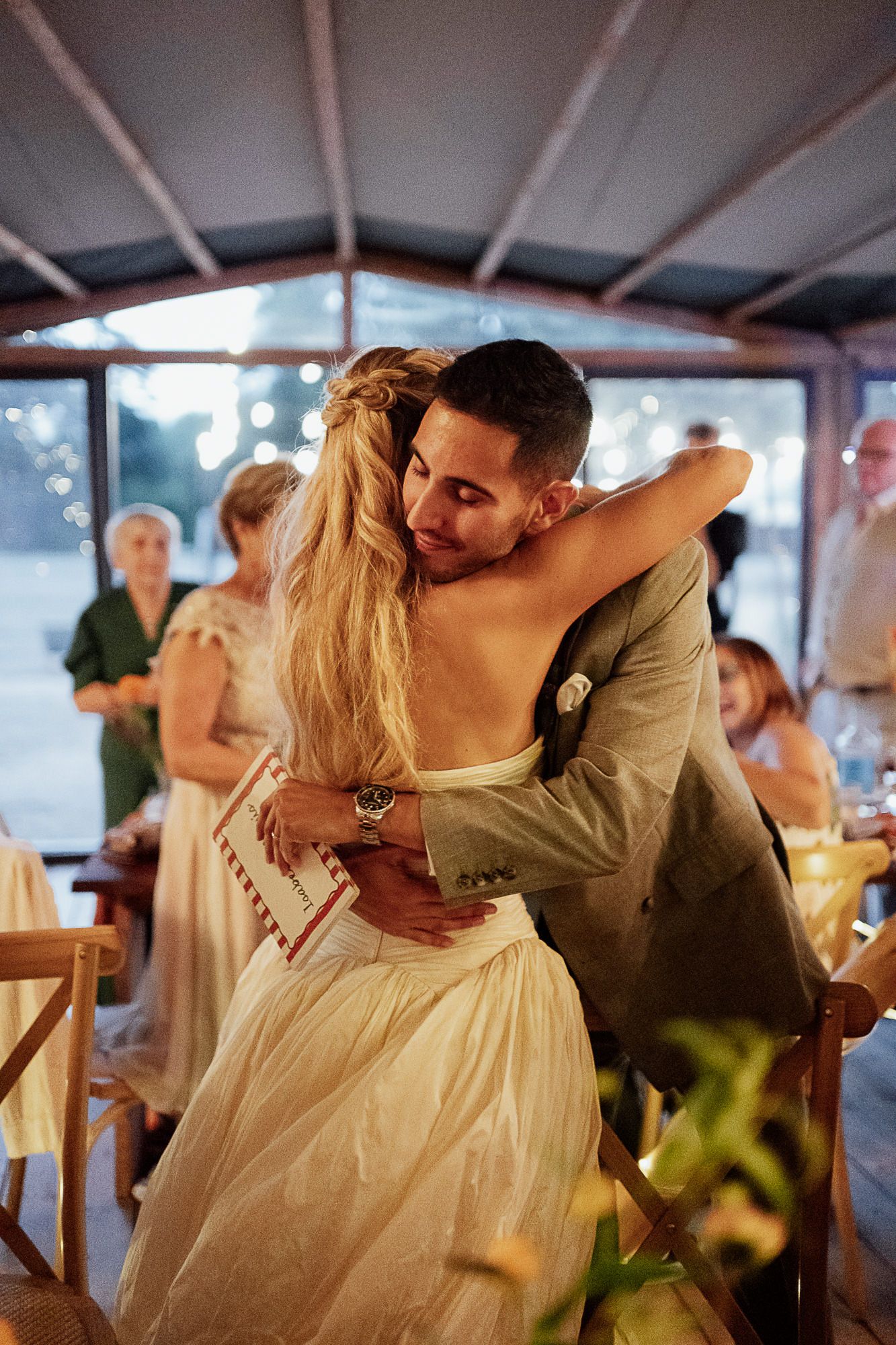 Bride and groom share an emotional hug during their wedding reception, surrounded by guests in a warmly lit venue.