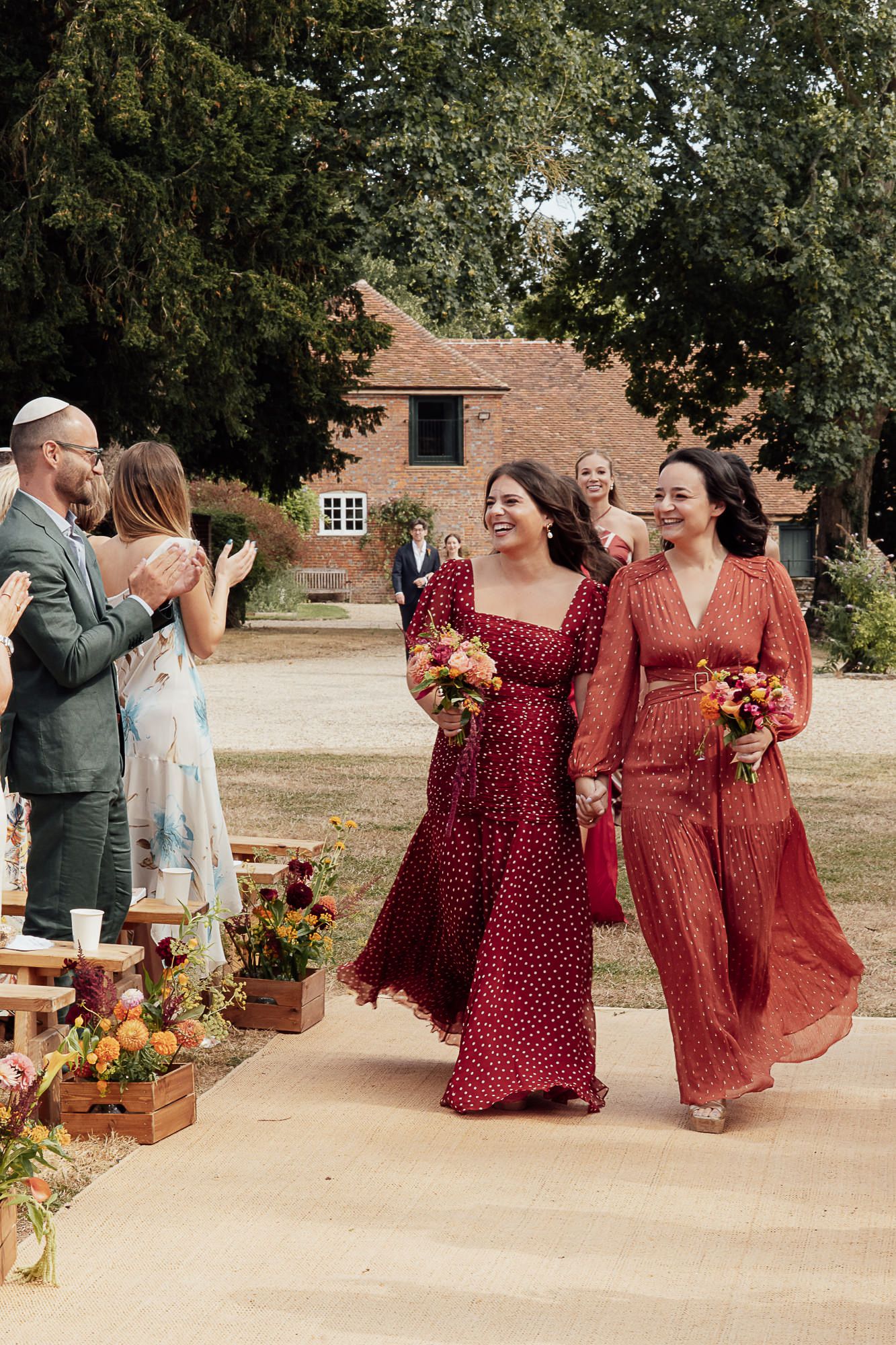 Two bridesmaids in red floral dresses walk down outdoor wedding aisle holding colorful bouquets at rustic venue.