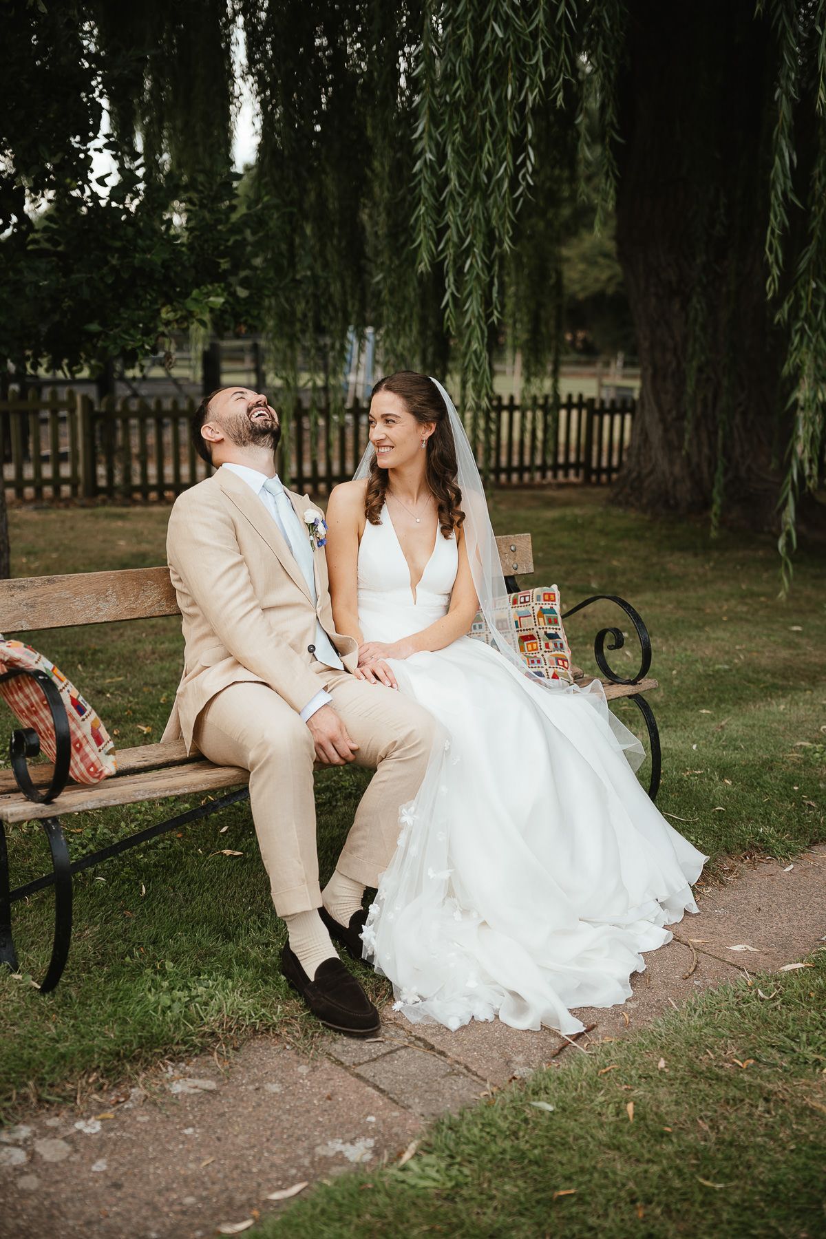 A couple in wedding attire sits together on a park bench under willow trees during an outdoor wedding photo session.