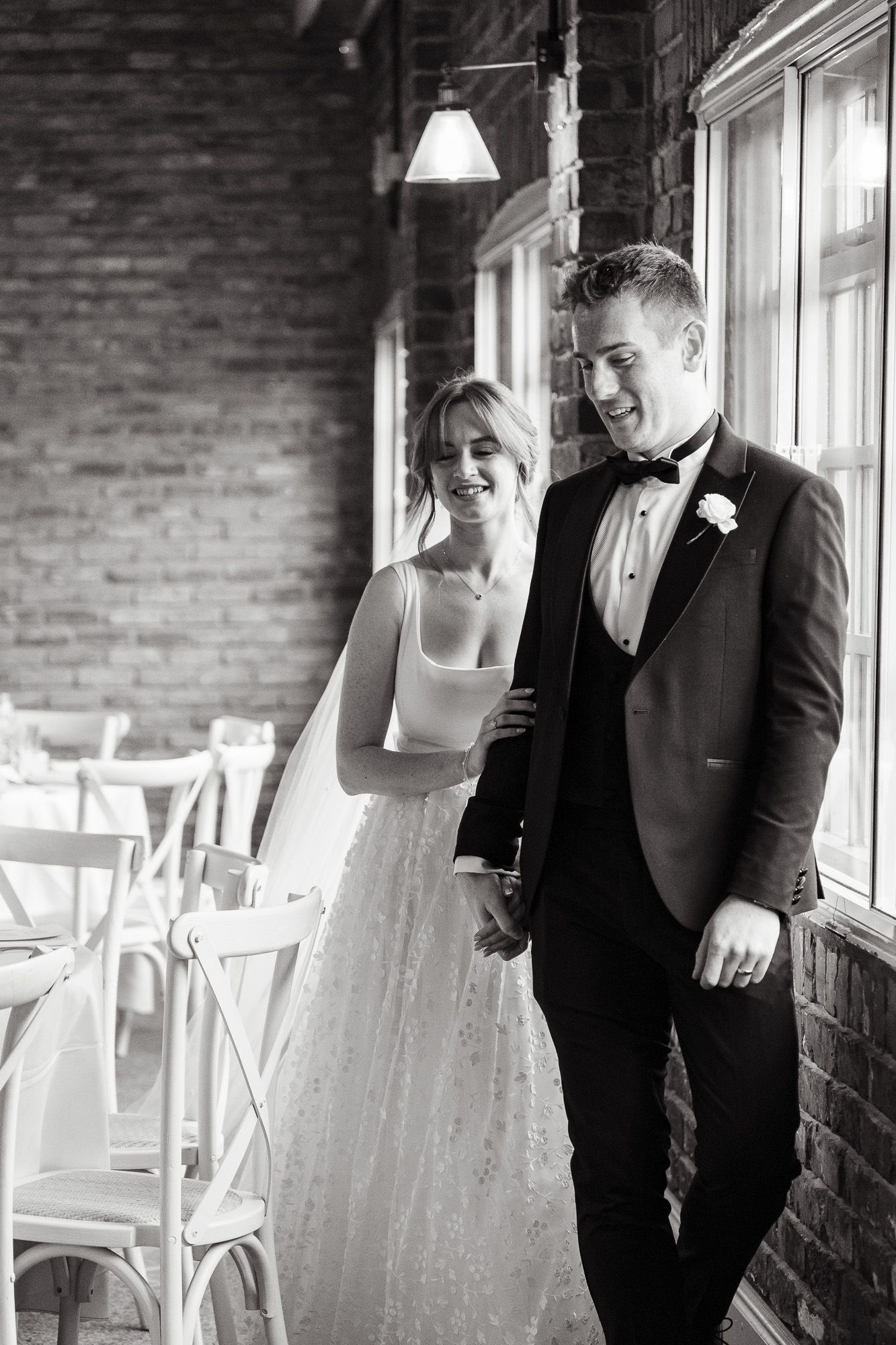A romantic black and white wedding photo of a couple standing together by a brick wall with elegant formal attire.