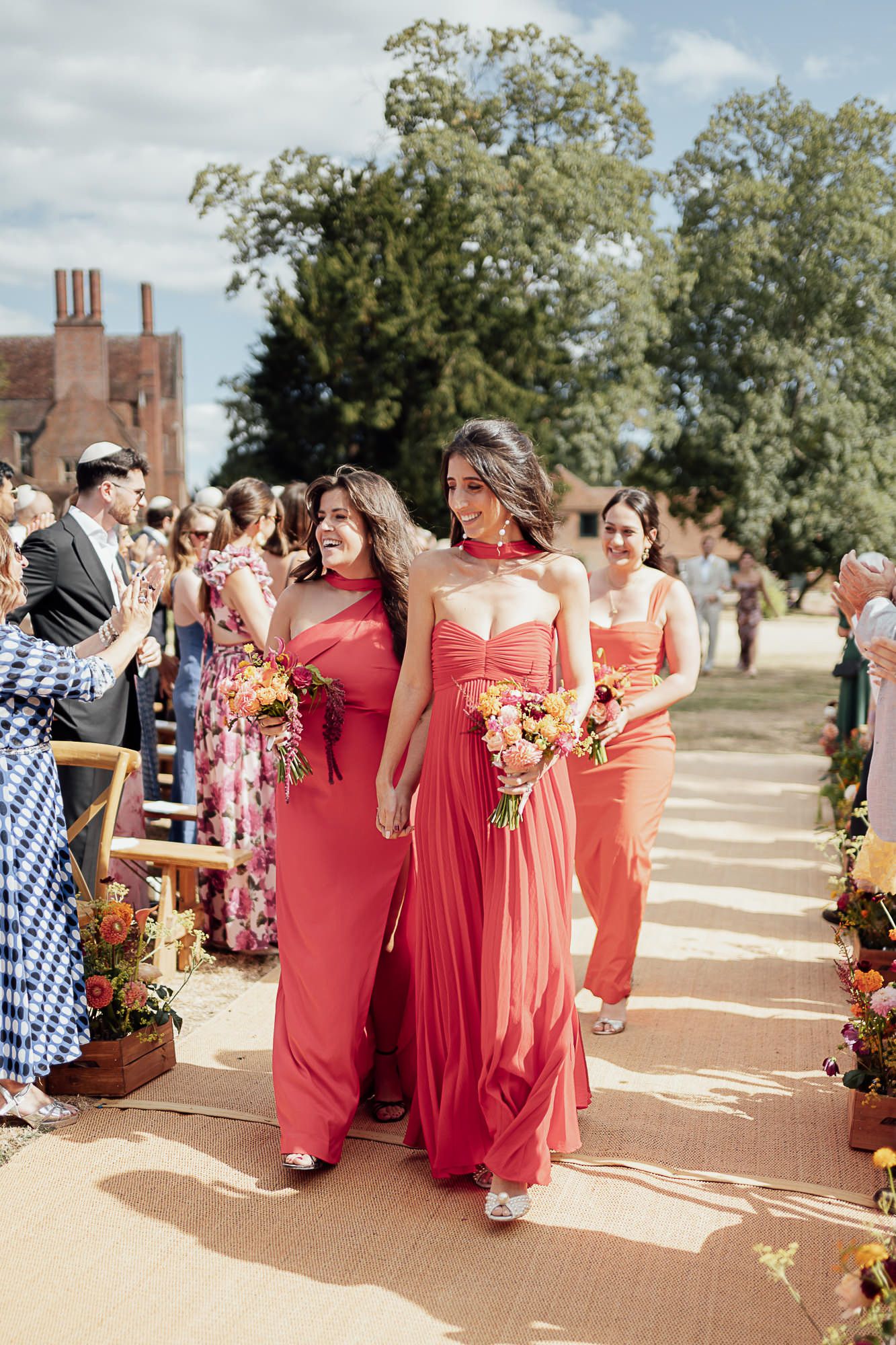 Bridesmaids in coral red gowns walk down outdoor wedding aisle holding colorful bouquets on a sunny day.