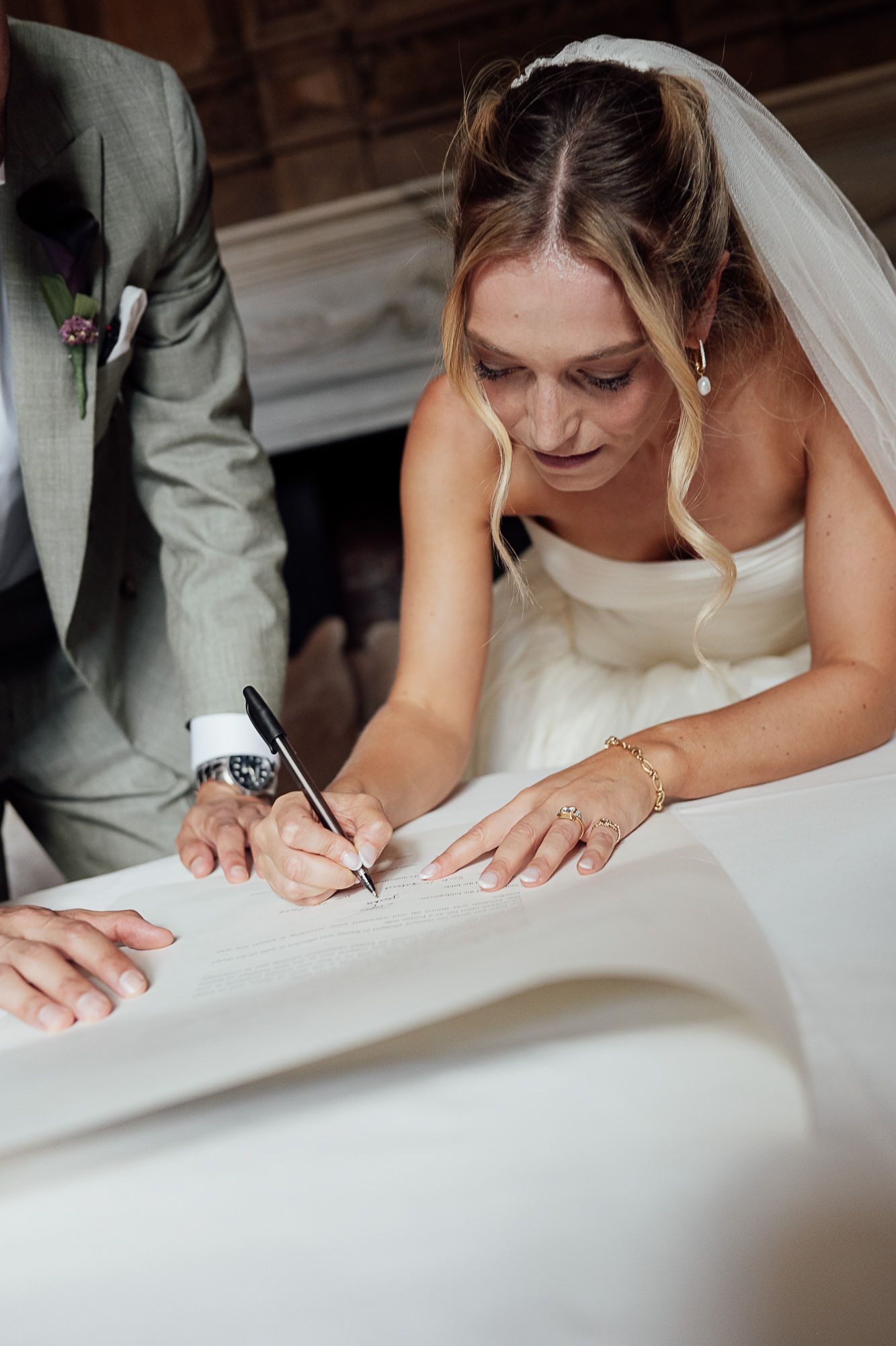 Bride in white dress and veil signing wedding certificate with pen at ceremony table.