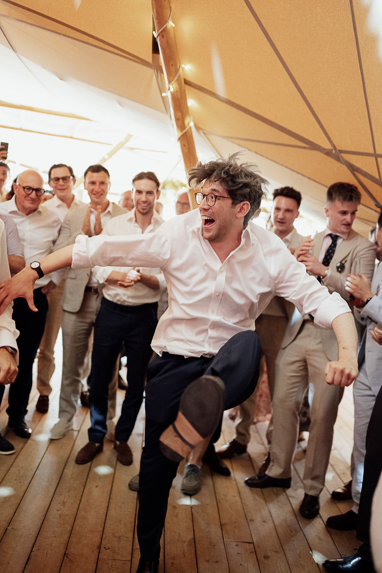 Energetic man dancing at a wedding reception in a marquee tent, surrounded by cheering guests in formal attire.