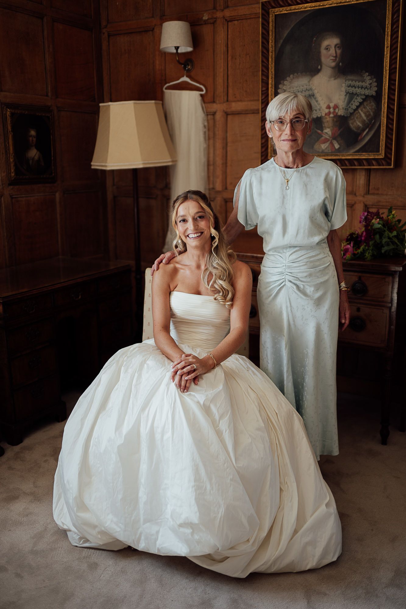 Bride in strapless ball gown sits beside elegant older woman in blue dress inside a wood-paneled room with portrait art.