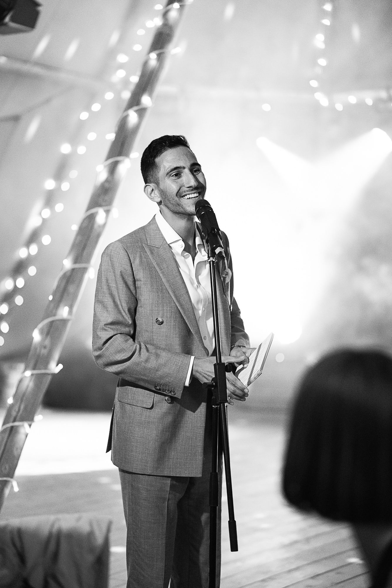 A smiling man in a suit speaks at a microphone stand at an elegant wedding reception with fairy lights.