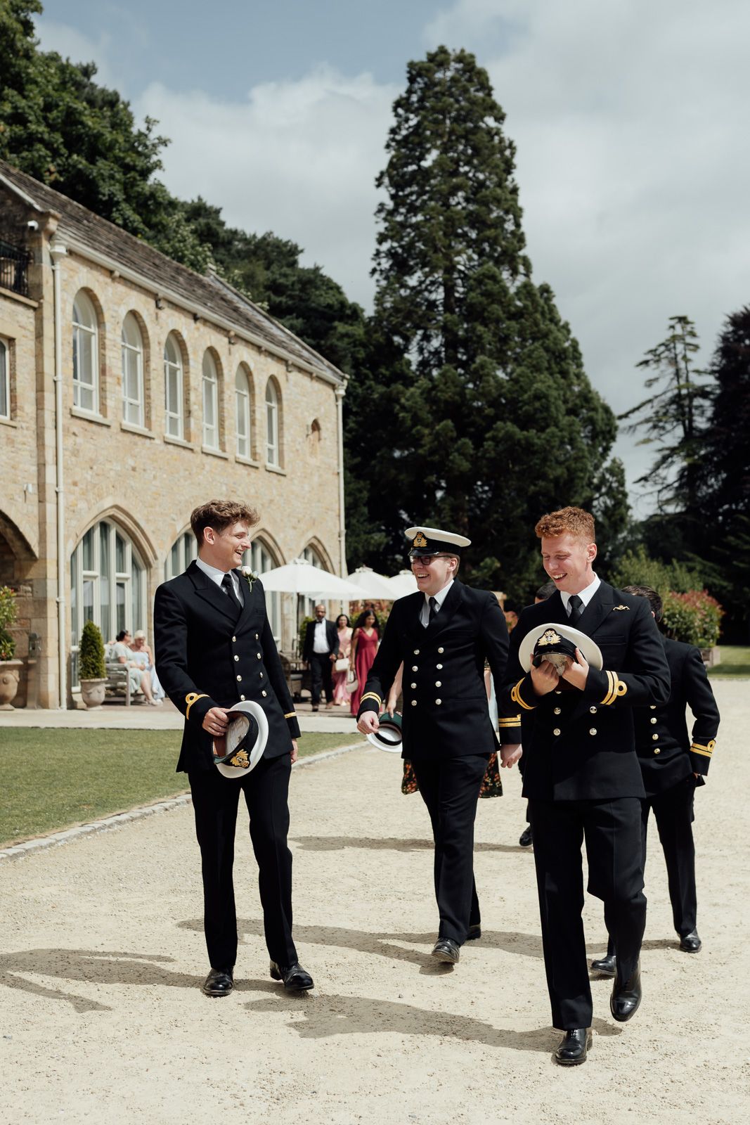 Three naval officers in dress uniforms walk along a gravel path outside a historic stone building on a sunny day.