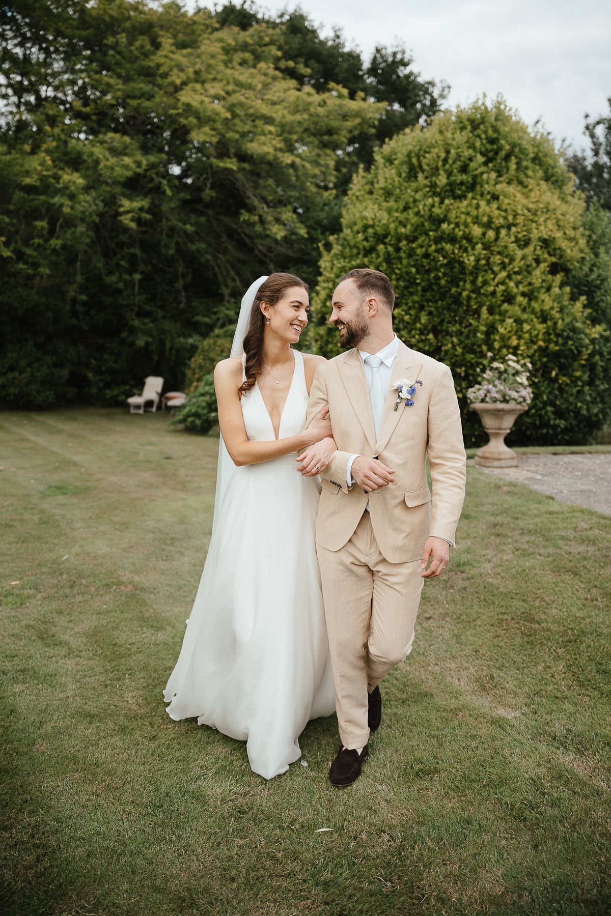 A couple in wedding attire walks together on a grassy lawn with lush greenery in the background.