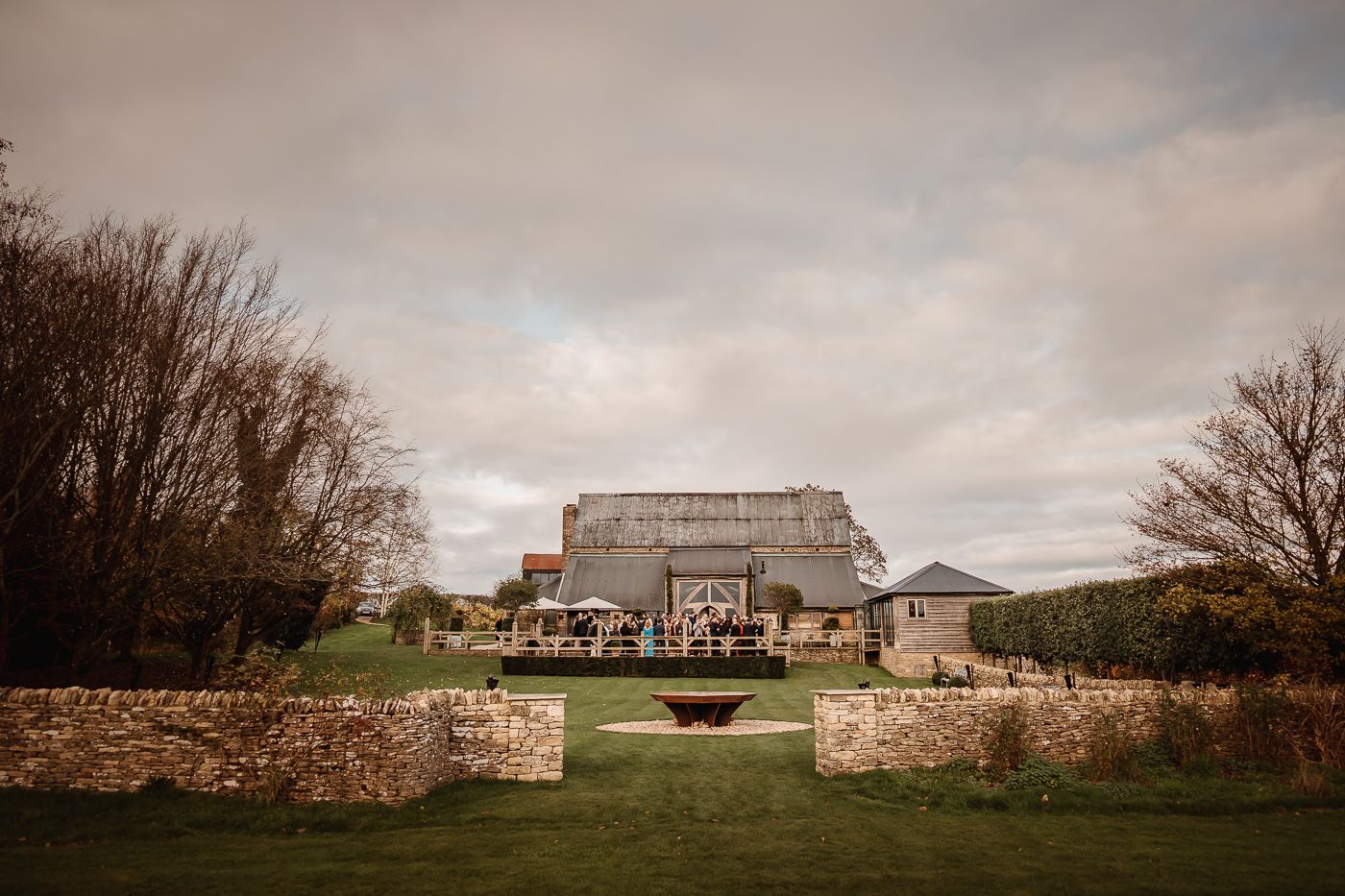 A rustic stone wall stretches across a rural landscape under cloudy skies with barns visible in the distance.