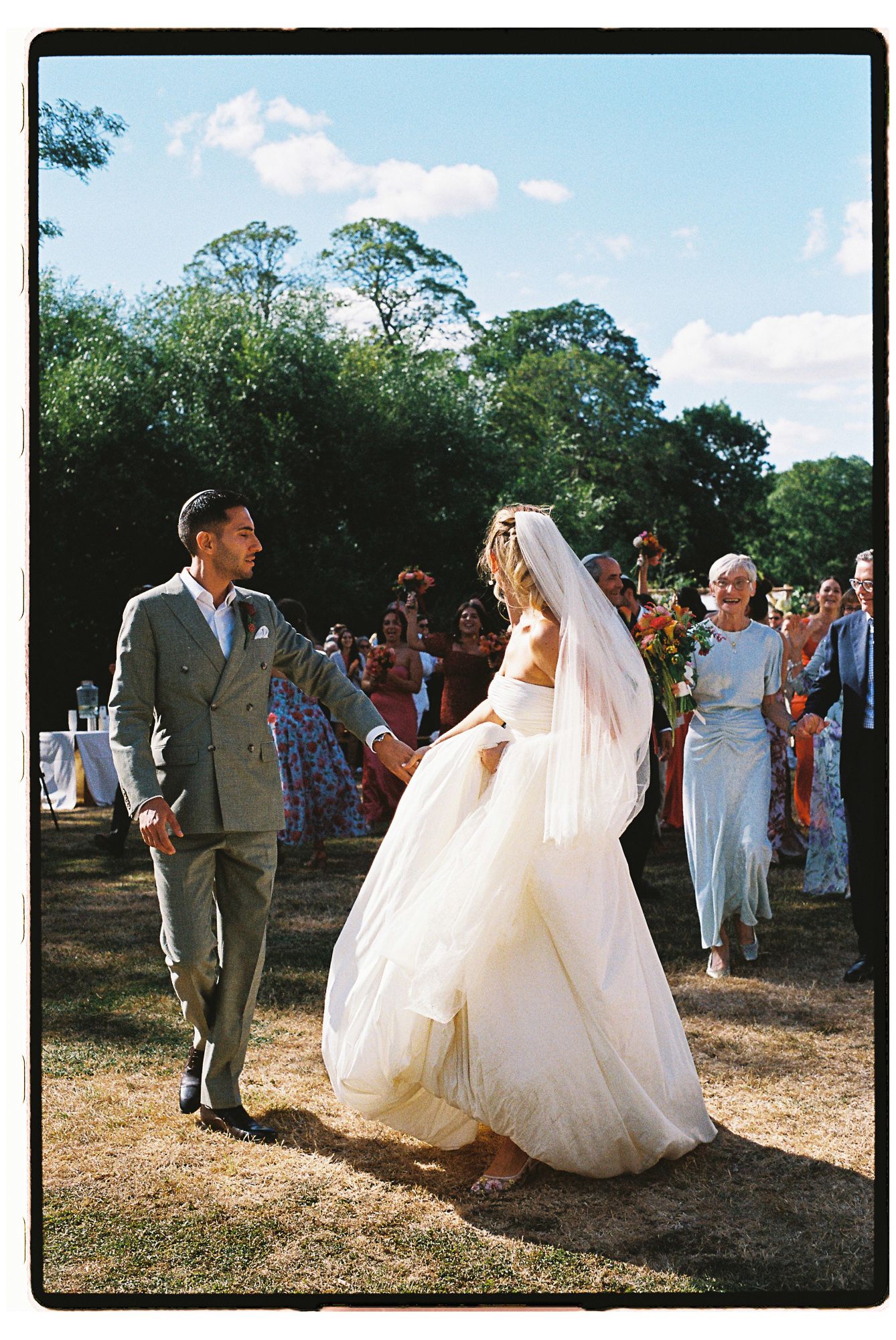Bride in flowing white gown and veil walks with groom in gray suit during outdoor wedding ceremony.