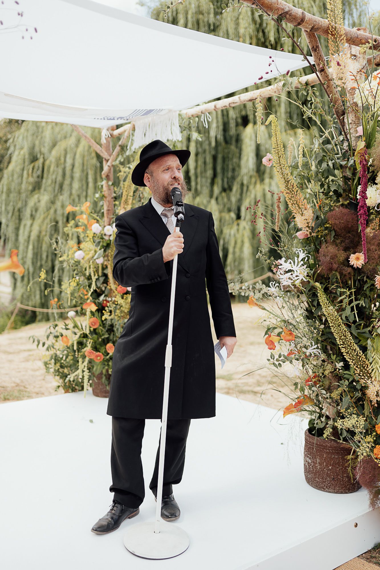 Rabbi in black coat and hat speaks at microphone during outdoor Jewish wedding ceremony.