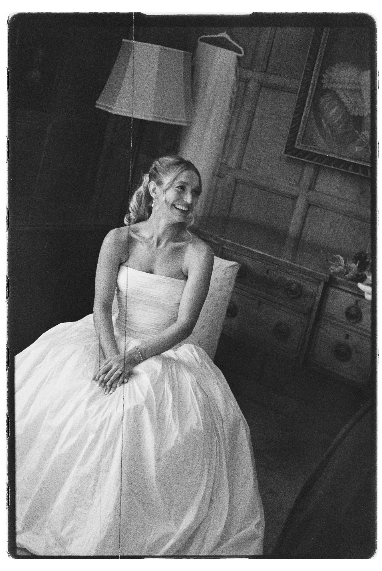 Black and white photo of a smiling bride in a strapless wedding gown seated in a room with a lamp and dresser.