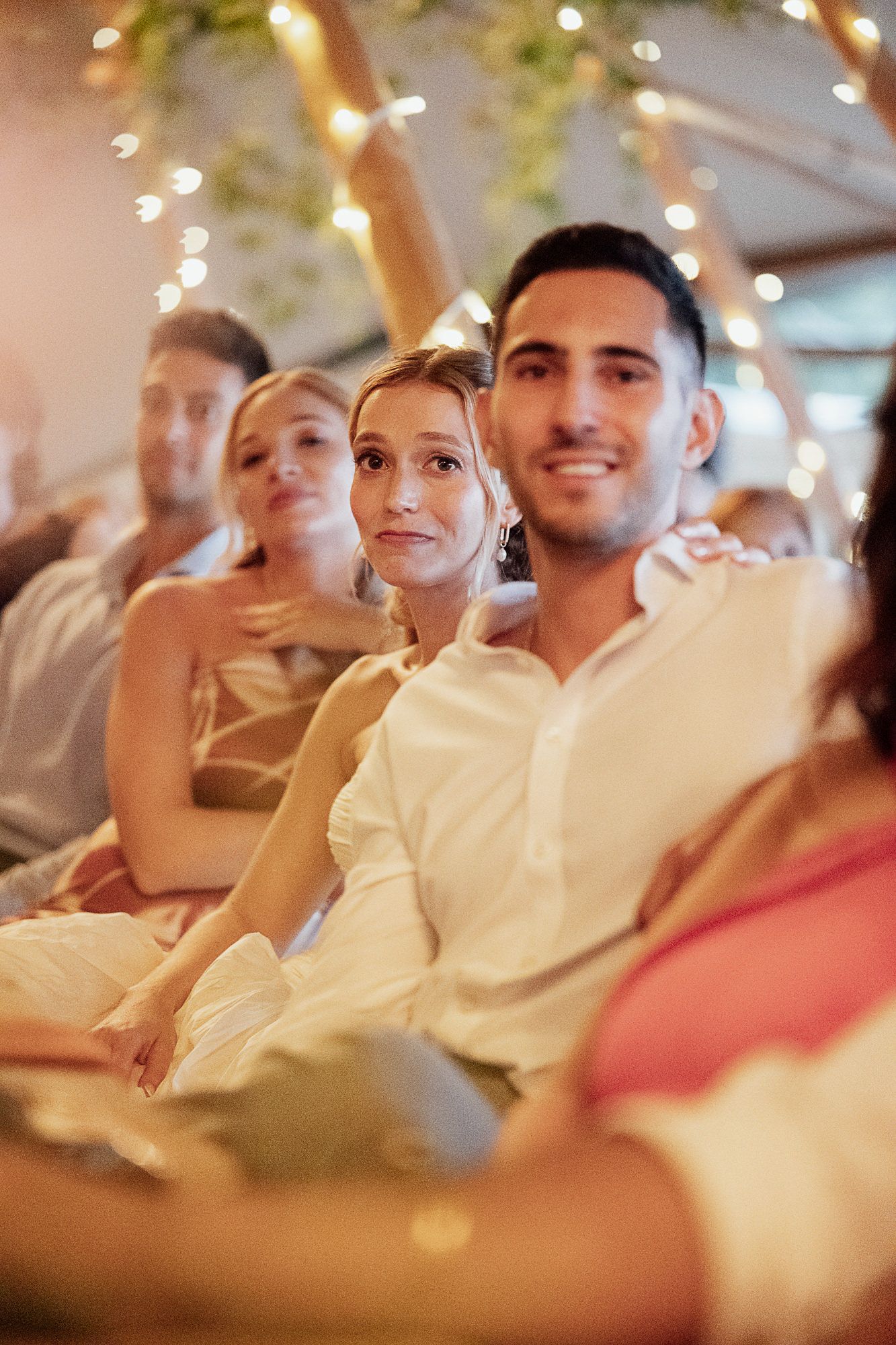 Smiling couple in white attire sitting together at a warmly lit wedding reception with guests in background.