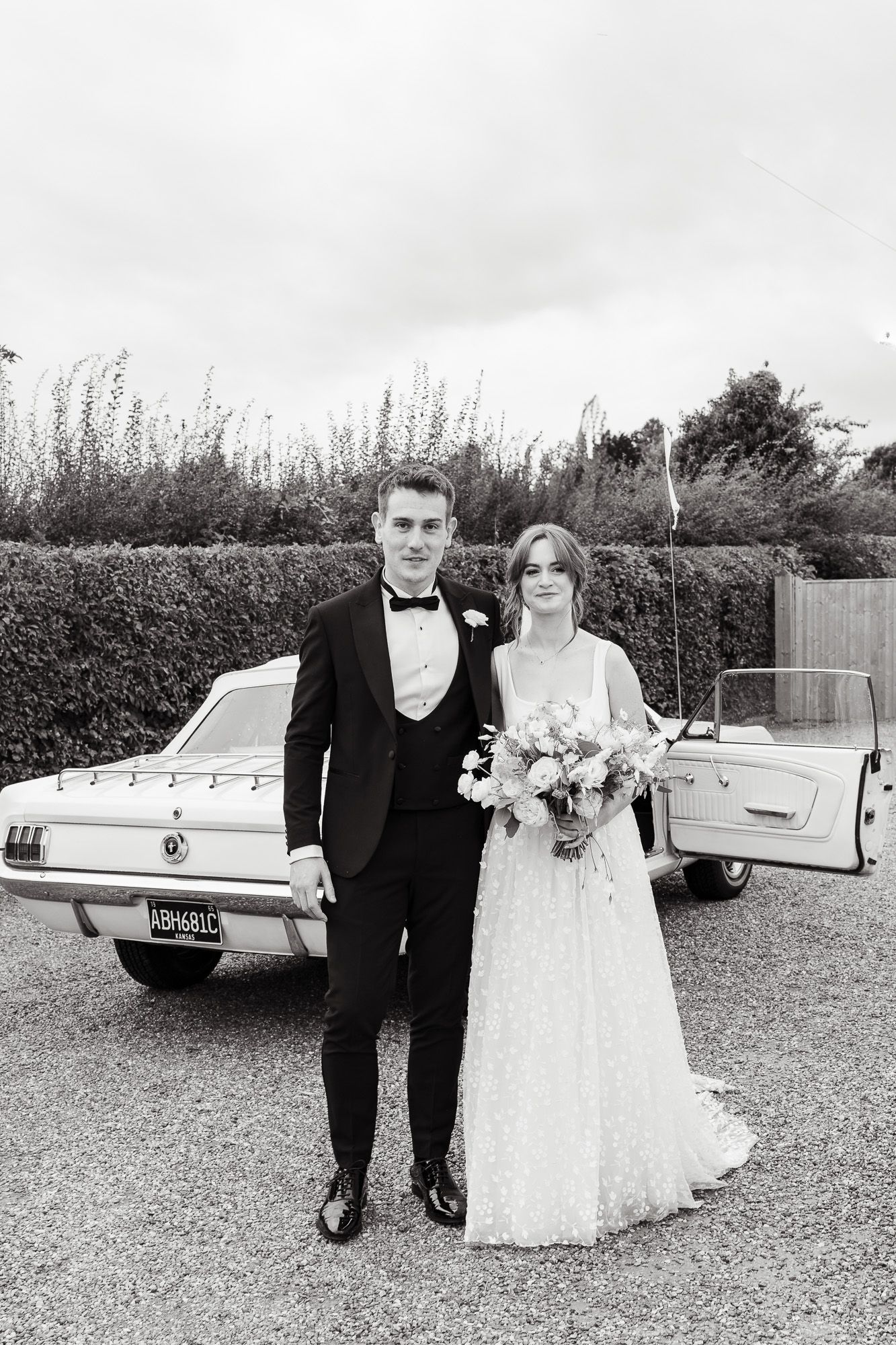 Black and white wedding portrait of a couple standing beside a vintage car on a gravel driveway.