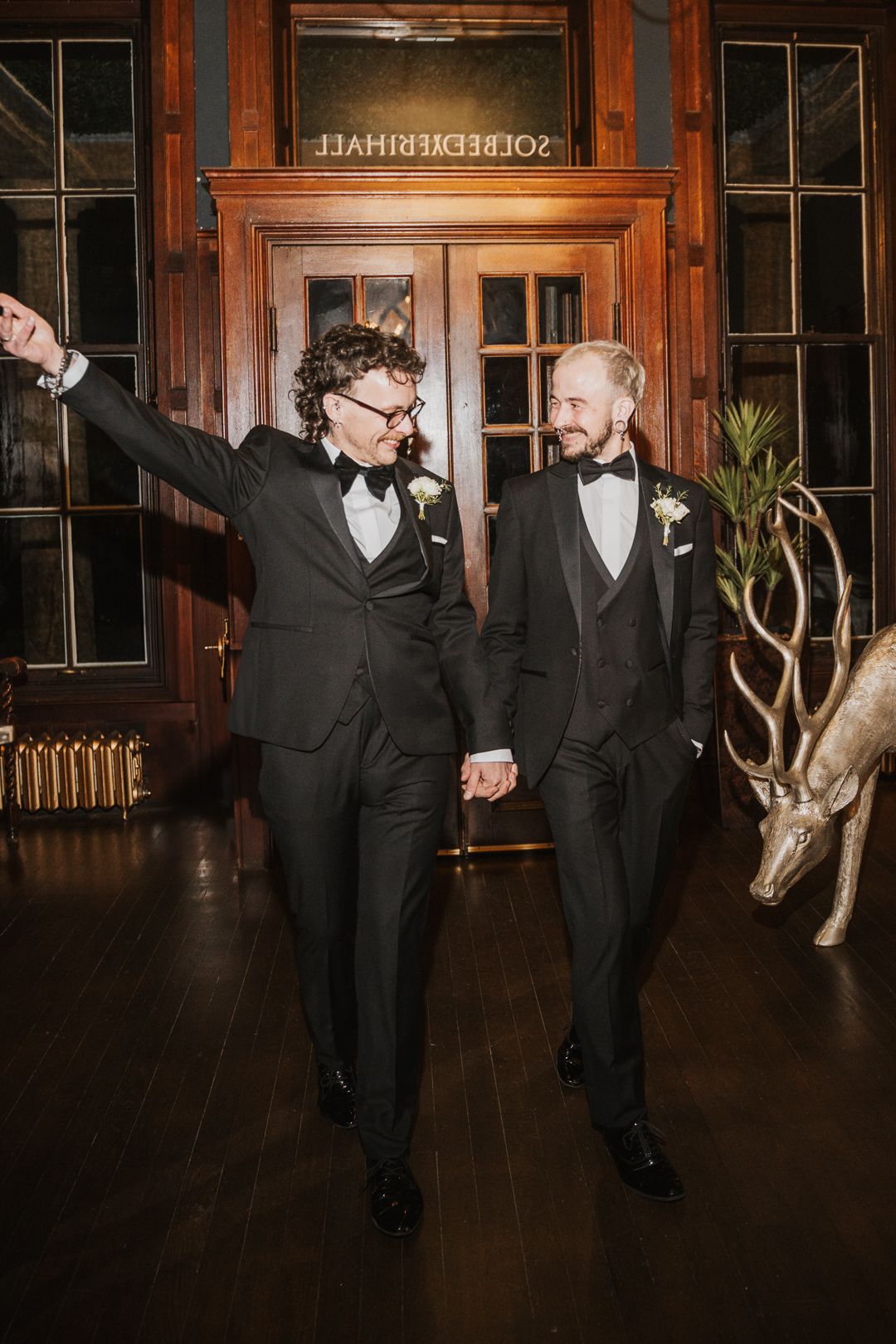 Two wedding guests in matching tuxedos dance and celebrate together in an elegant venue with wood paneling.