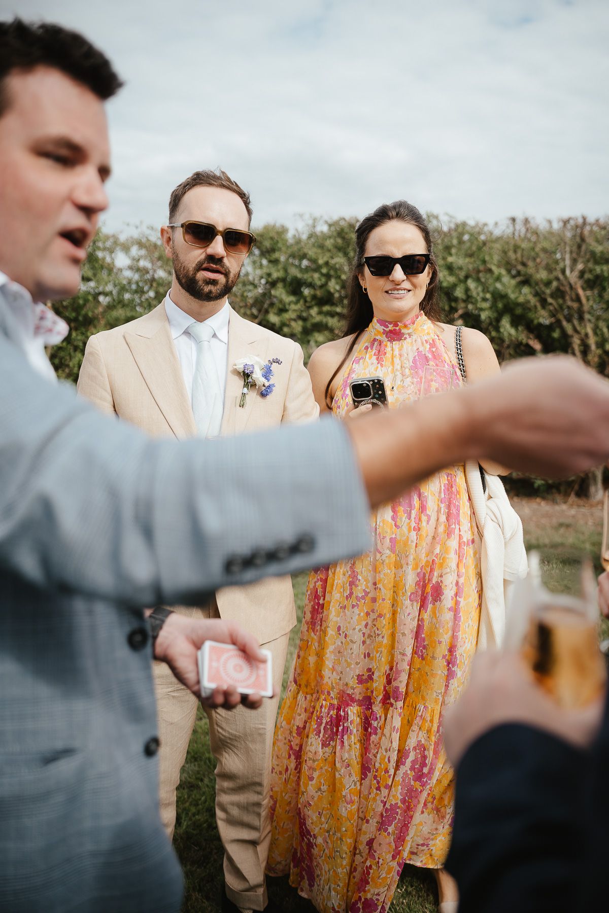 Wedding guests in formal attire gather outdoors, with someone wearing a peach-colored suit and another in a floral summer dress.