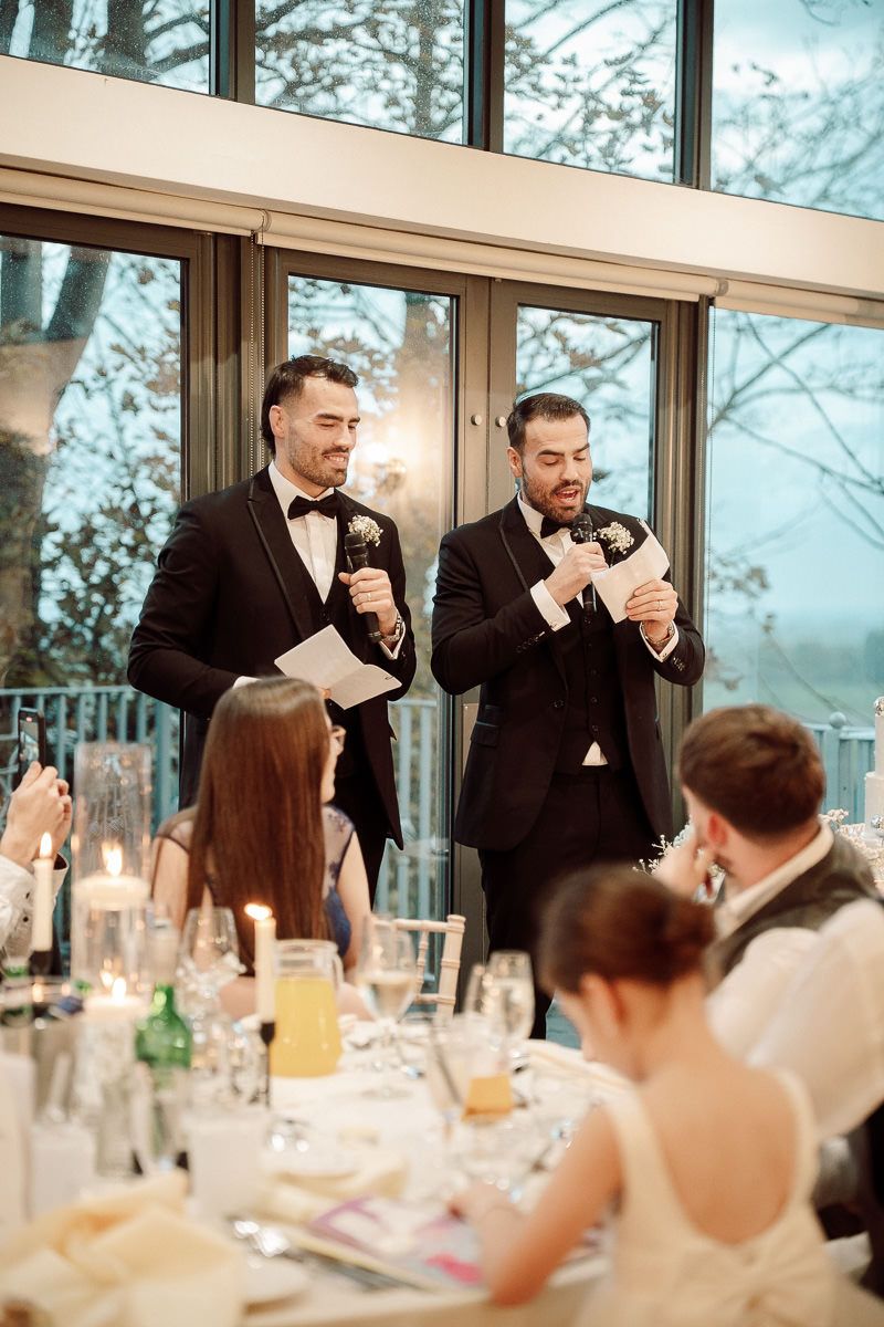 Two groomsmen in black tuxedos giving wedding speeches at an elegant reception dinner.