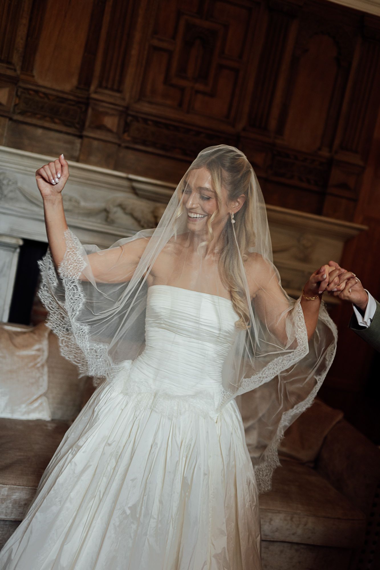 Joyful bride in strapless white gown and lace veil dancing with arms raised in ornate wedding venue.