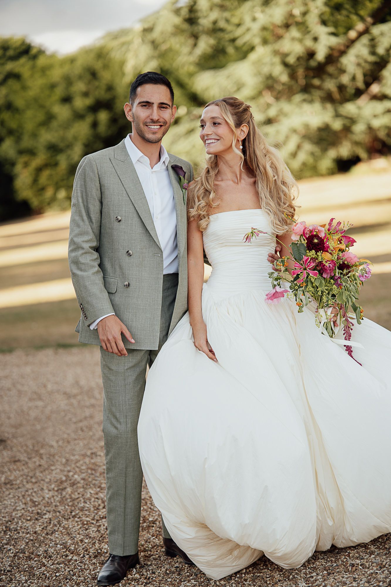 A smiling bride in a strapless white ballgown holds a vibrant bouquet beside a groom in a grey suit outdoors.