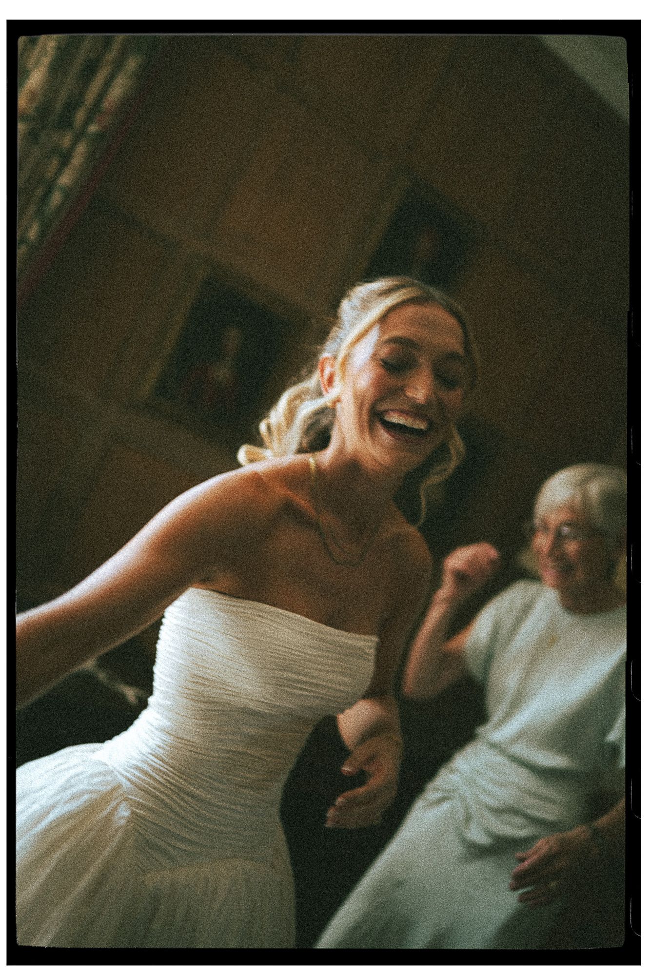 A joyful bride in a strapless white gown laughs and dances while an elderly woman smiles in the background.