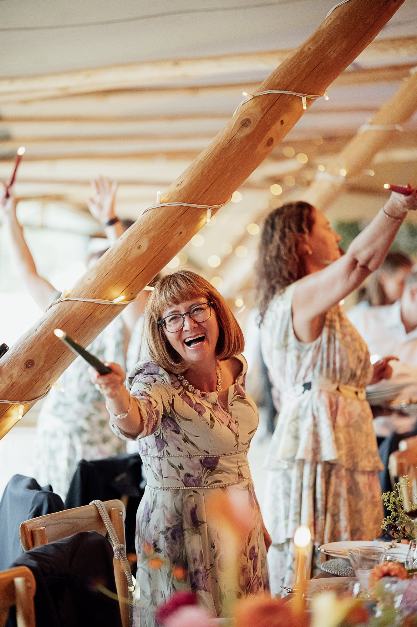 Happy woman in floral dress laughing and dancing with raised arms at a festive wedding reception.